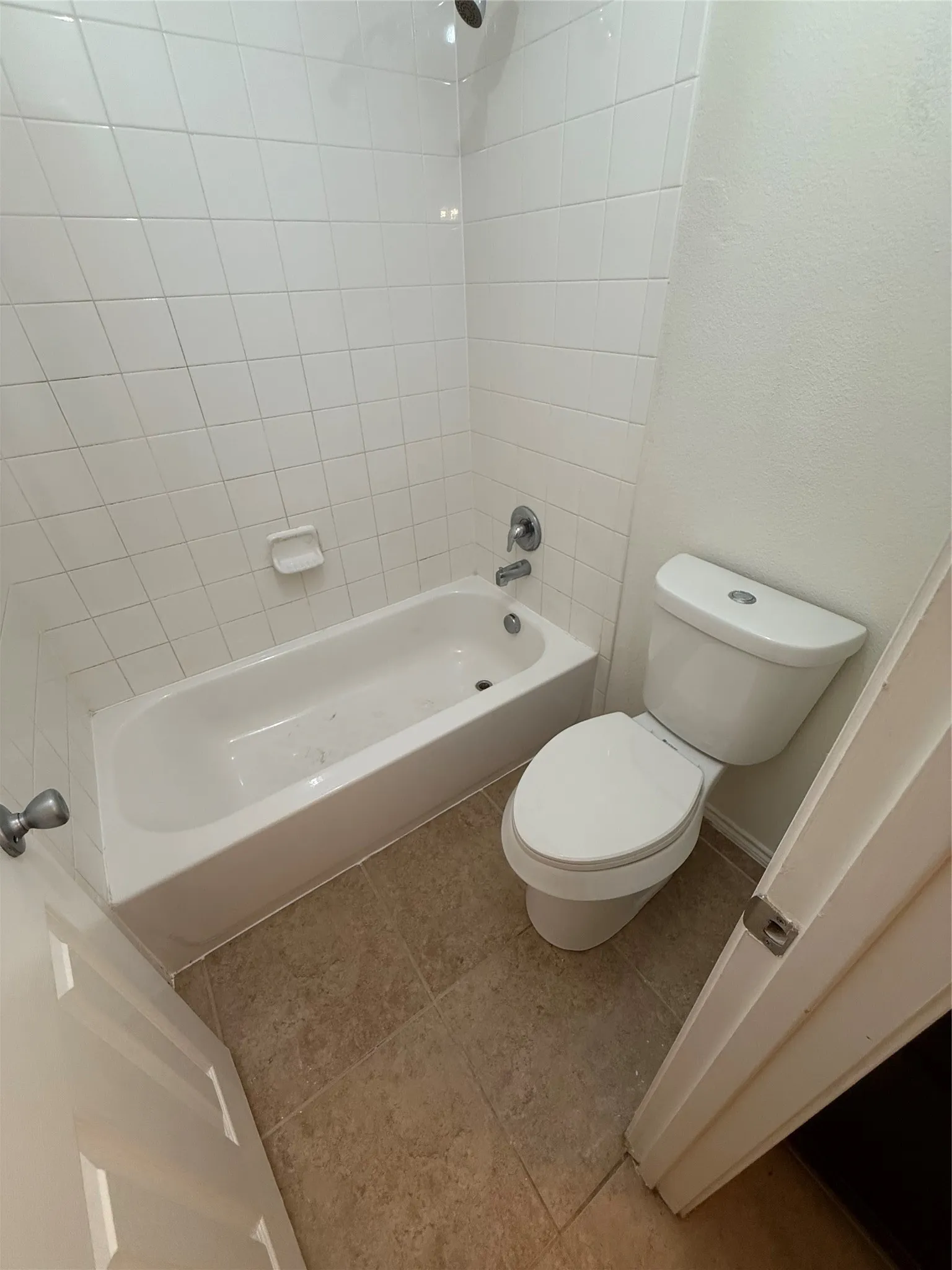 White tiled bath and shower combination with a chrome showerhead and fixtures, alongside a white toilet, set on a tan stone-look tile floor