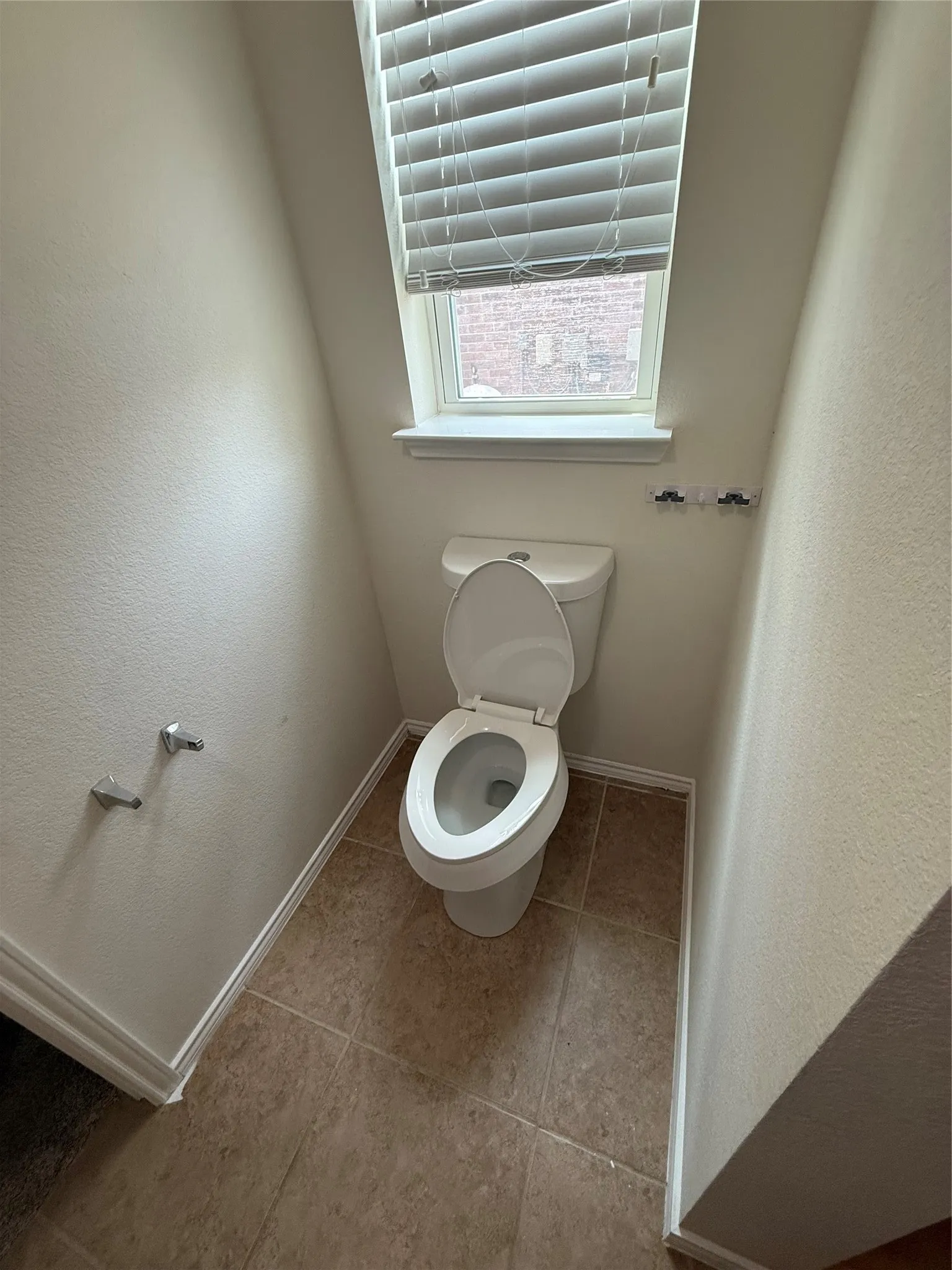 White toilet with a window, white window trim, and horizontal blinds set above tan tile flooring