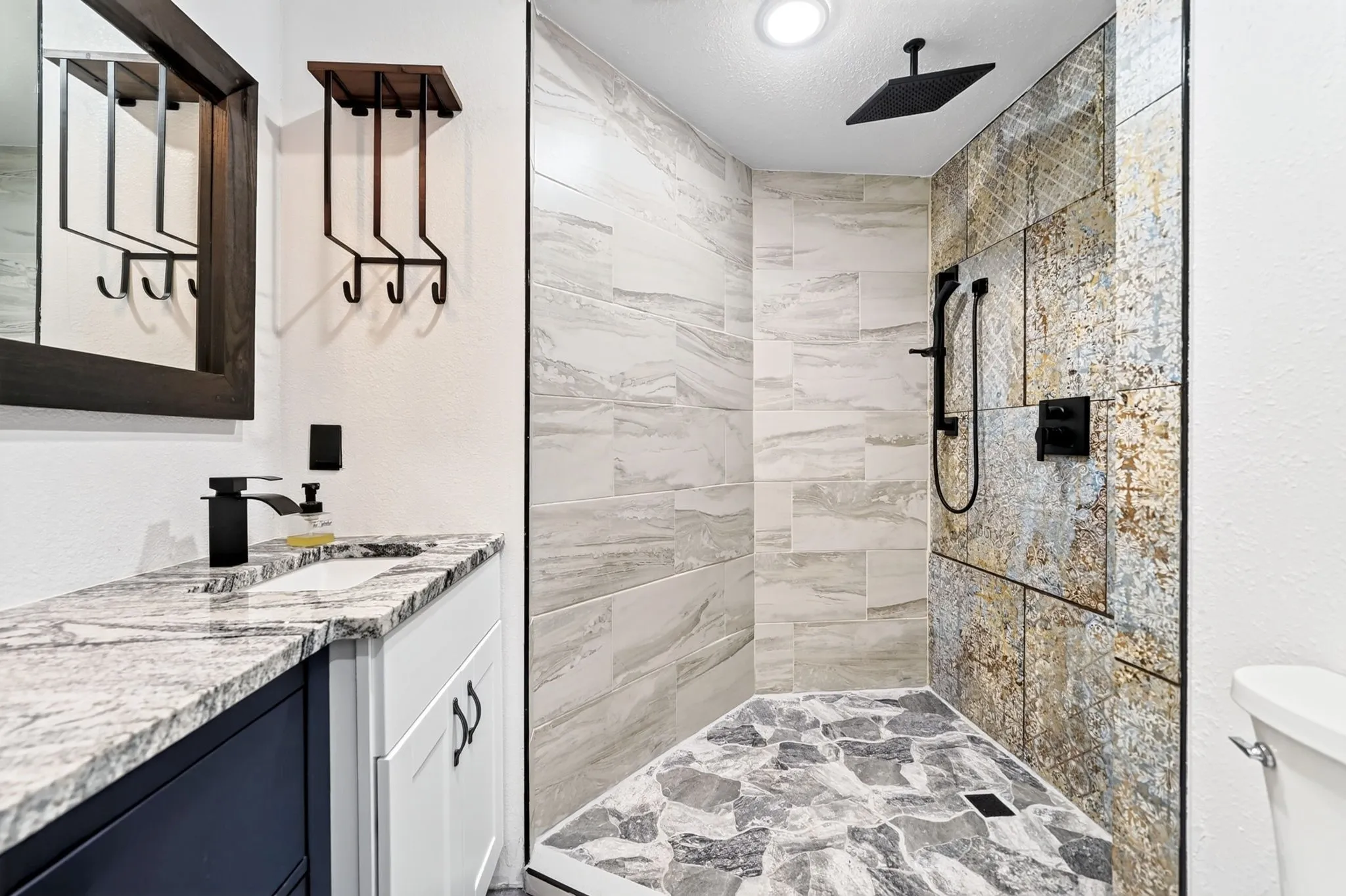 Shower with patterned wall tile and rainfall showerhead, stone-finish shower pan, vanity with a rectangular sink, matte black faucet, and granite countertop