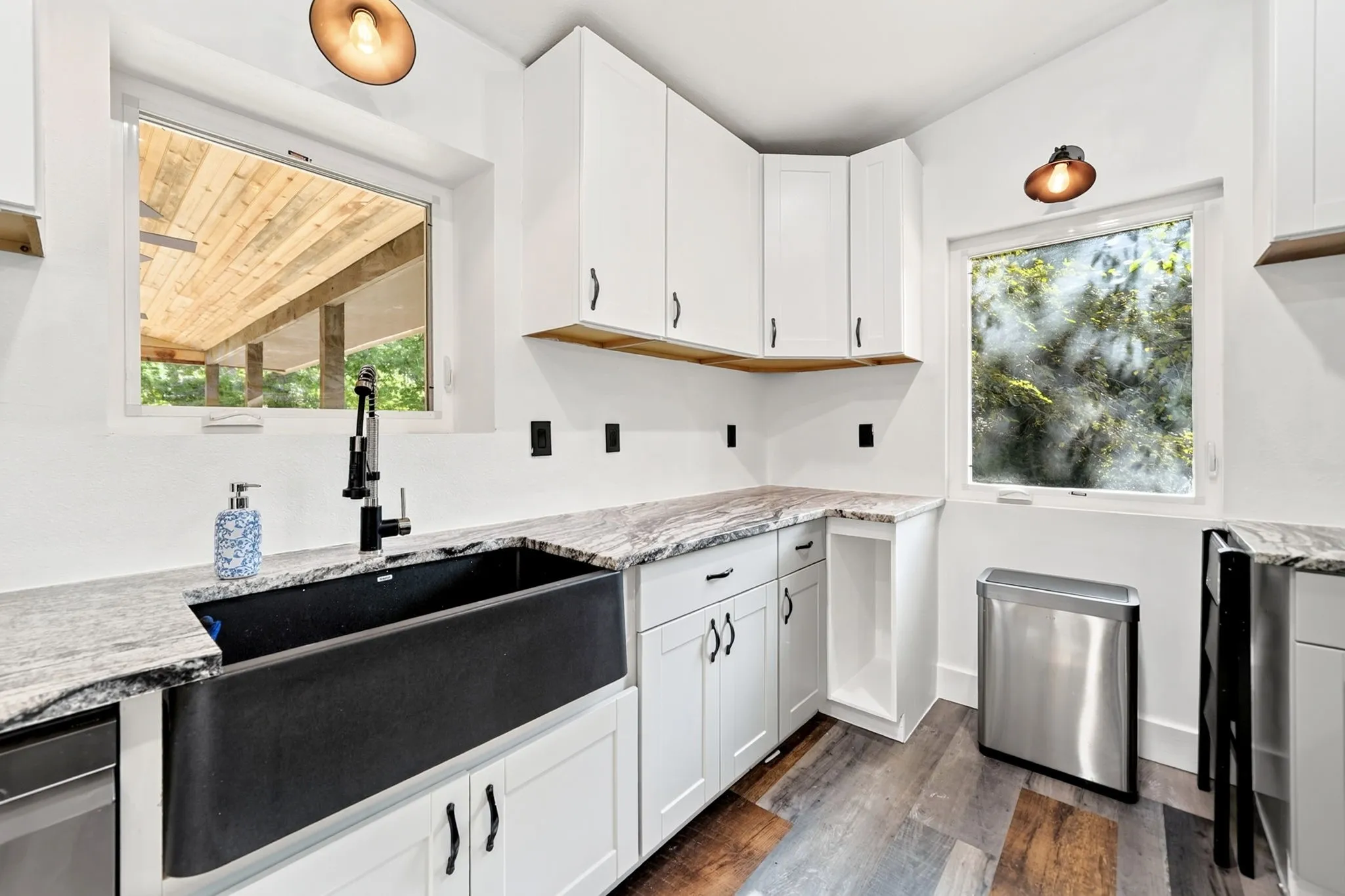 Modern kitchen featuring white cabinetry, stone countertops, wood-finish flooring, a black farmhouse-style sink, and contemporary light fixtures