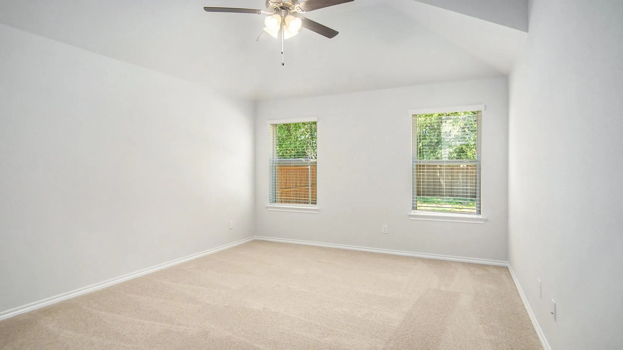 Spacious room featuring vaulted ceilings, light neutral carpeting, two windows with blinds, and a ceiling fan with integrated lighting