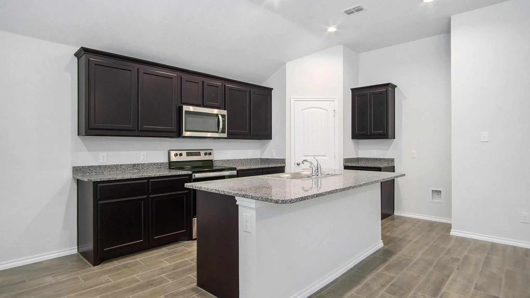 Modern kitchen featuring a central island with a sink, dark wood cabinetry, stainless steel appliances, granite countertops, and wood-finish flooring