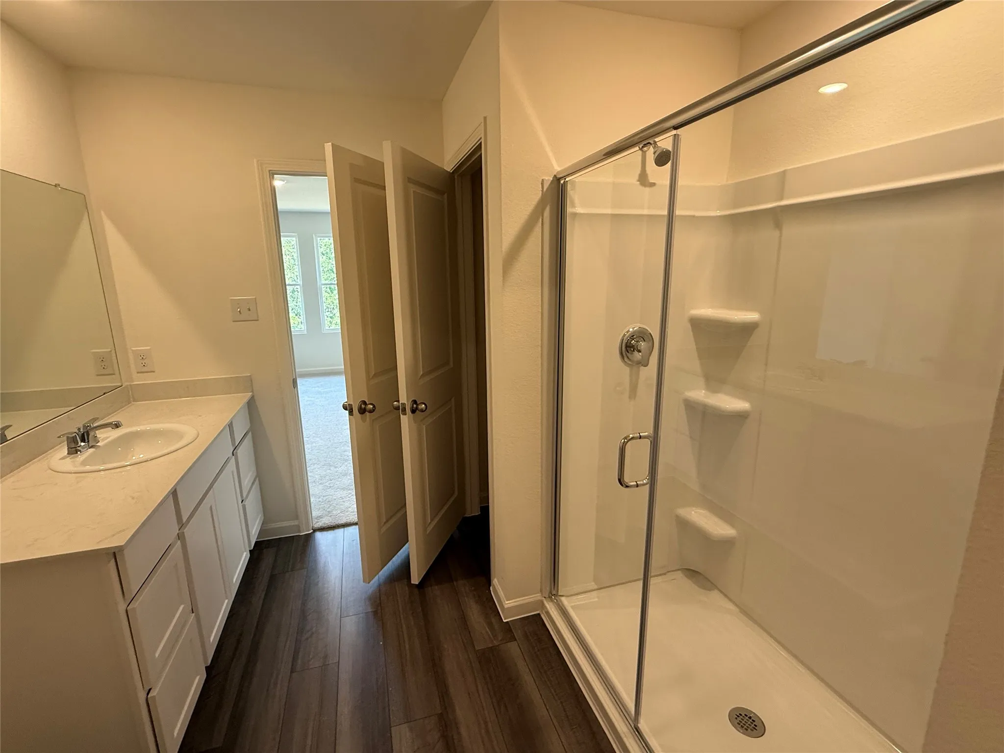 Wood-finish flooring, single vanity with light-colored countertop, chrome faucet, and glass-enclosed shower with built-in shelving