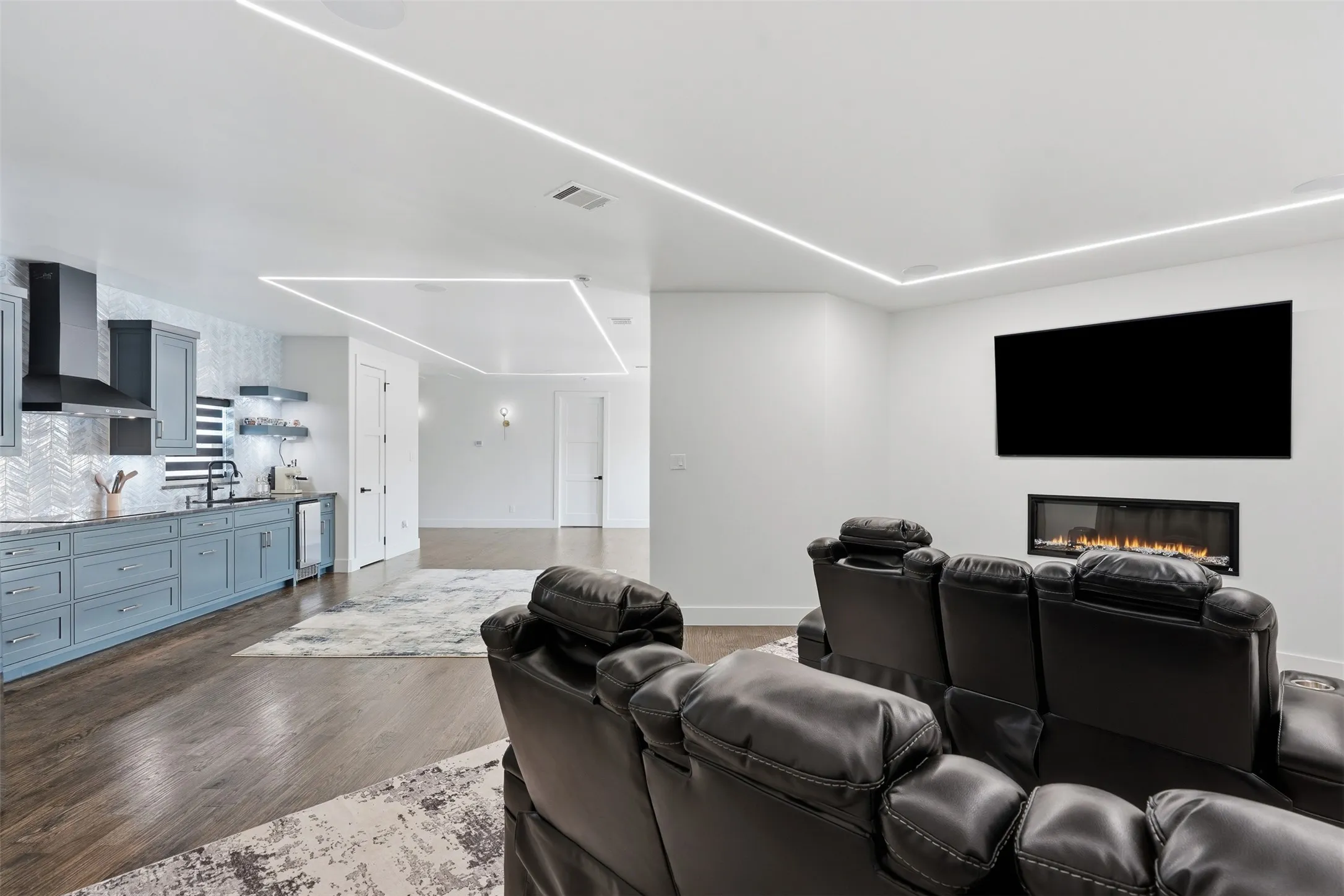 Open-concept living space featuring wood-finish flooring, linear ceiling lighting, a built-in electric fireplace, and a kitchen with blue cabinetry and a tiled backsplash