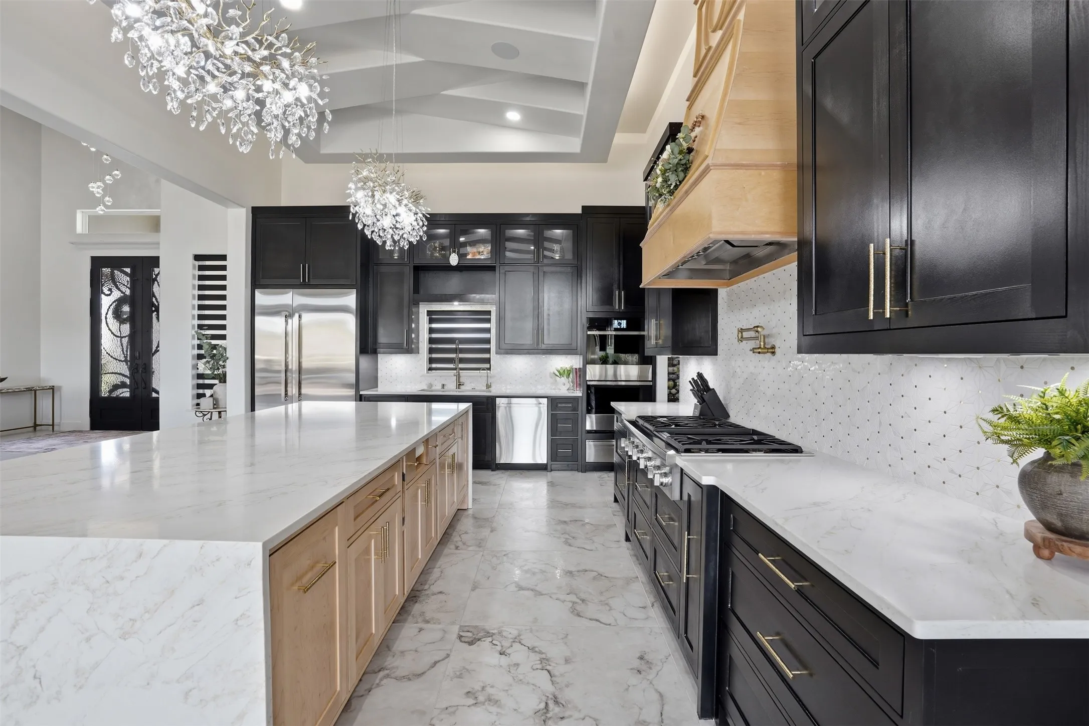 Gourmet kitchen featuring dual islands with white stone countertops, black cabinetry with brushed gold hardware, stainless steel appliances including a gas cooktop, white subway tile backsplash, polished stone flooring, and statement crystal chandelier lighting