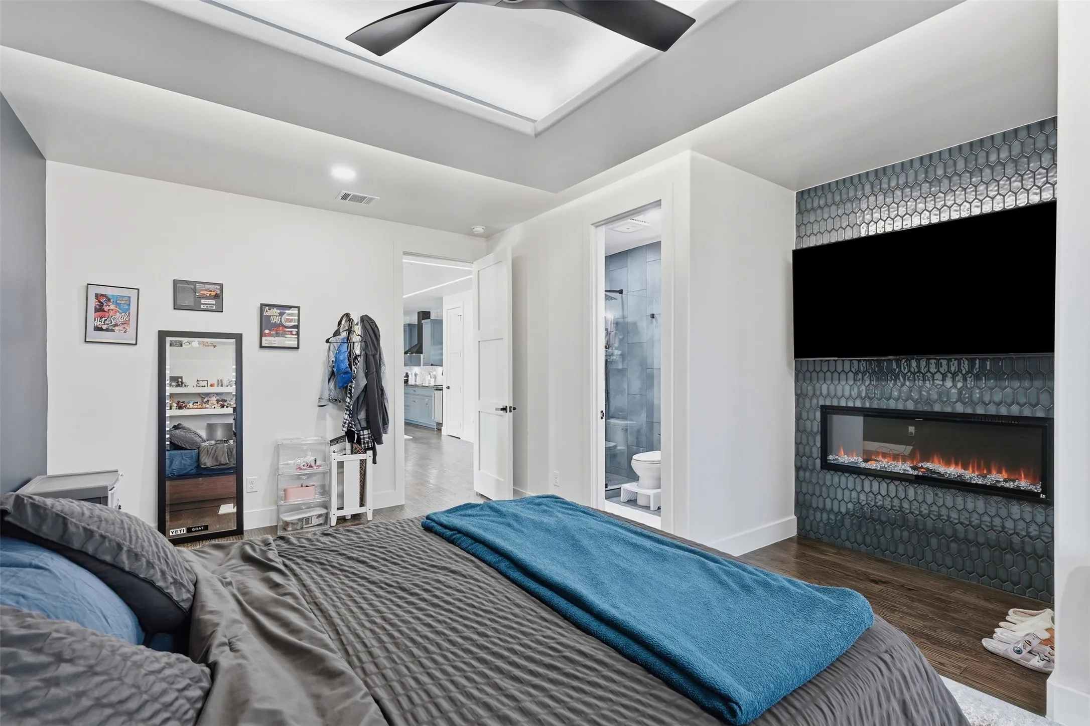 Master bedroom featuring a coffered ceiling with integrated recessed lighting and ceiling fan, wood-finish flooring, and an ensuite with linear fireplace, tile accents, and flat-screen television
