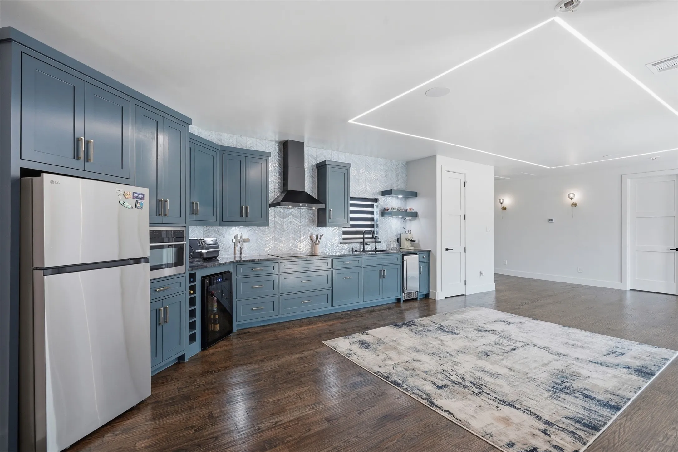 Modern kitchen and open living area featuring custom cabinetry in slate blue with stainless steel appliances, white subway tile backsplash, wood-finish flooring throughout, recessed and linear ceiling lighting, and seamless integration with the adjacent dining and living spaces