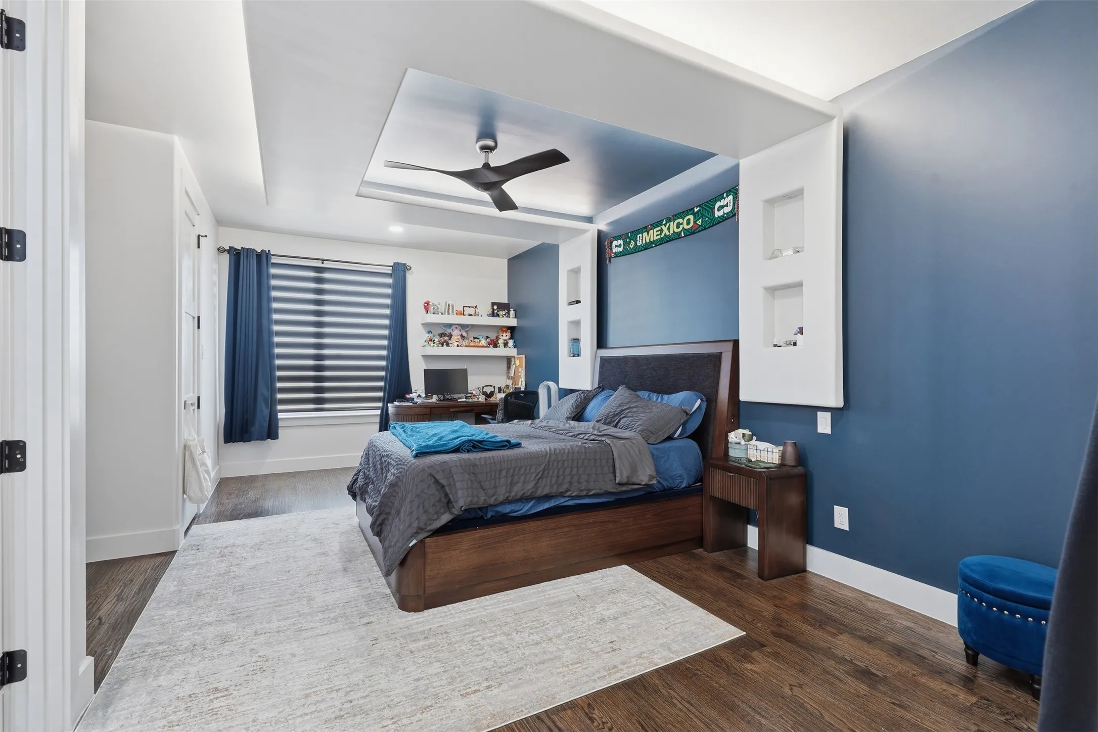 Recessed ceiling with a contemporary ceiling fan, built-in shelving, wood-finish flooring, and a window with blinds