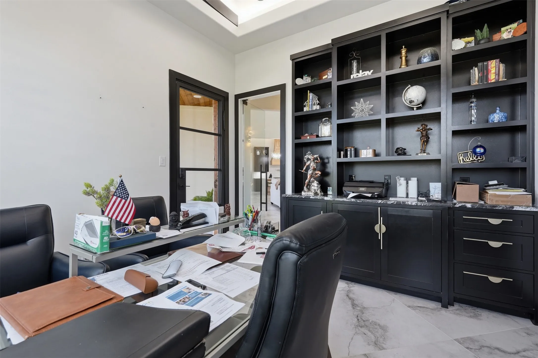 Executive office featuring floor-to-ceiling black cabinetry with glass-front shelving and lower storage drawers, polished stone-finish flooring, recessed ceiling lighting, and French doors with gridded glass panes opening to an adjacent room