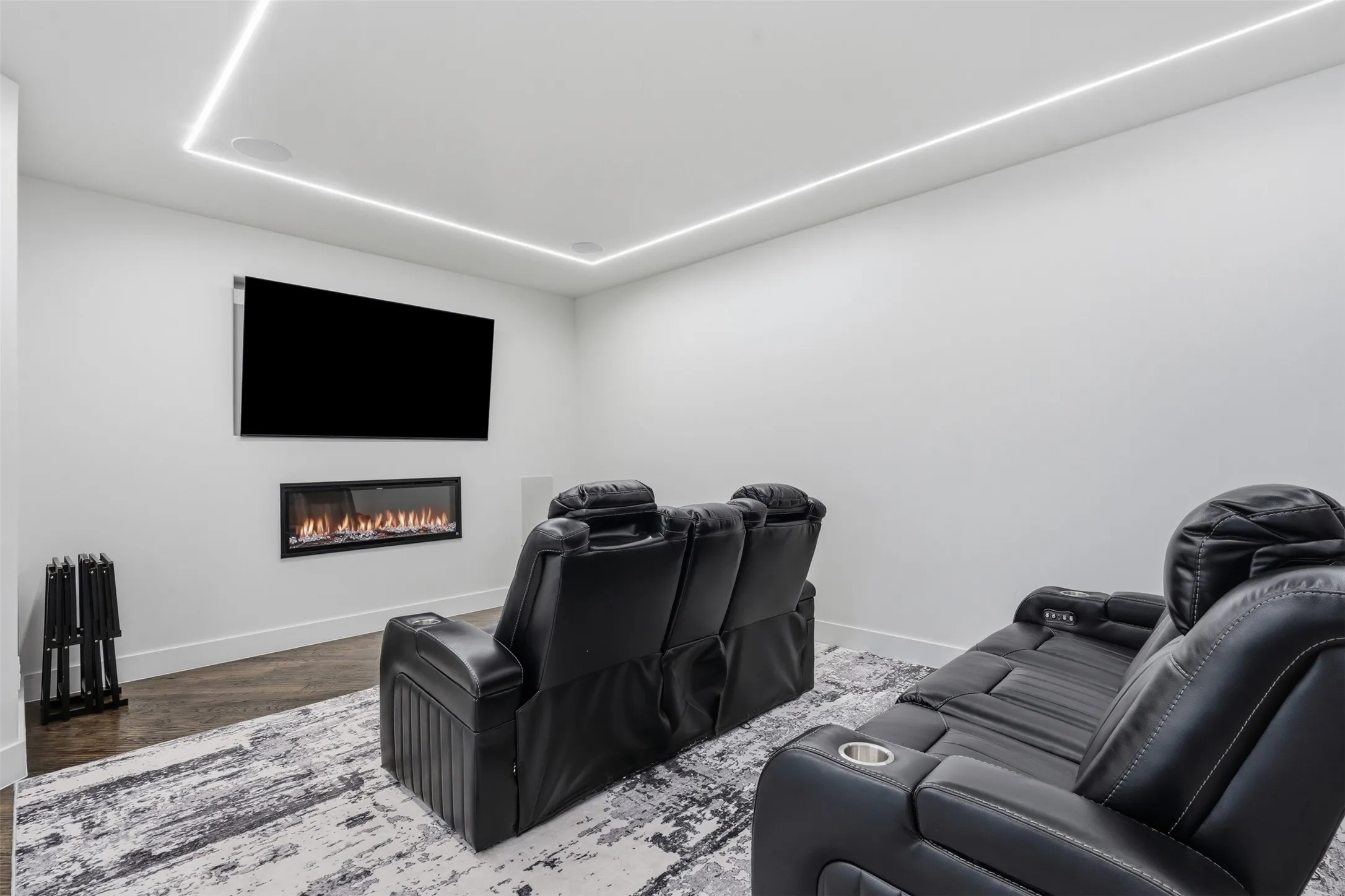 Home Theater Room featuring a wall-mounted flat-screen display, linear electric fireplace insert, premium black leather seating with integrated recliners and cup holders, geometric patterned area rug, and ambient ceiling-mounted LED lighting with architectural tray detail