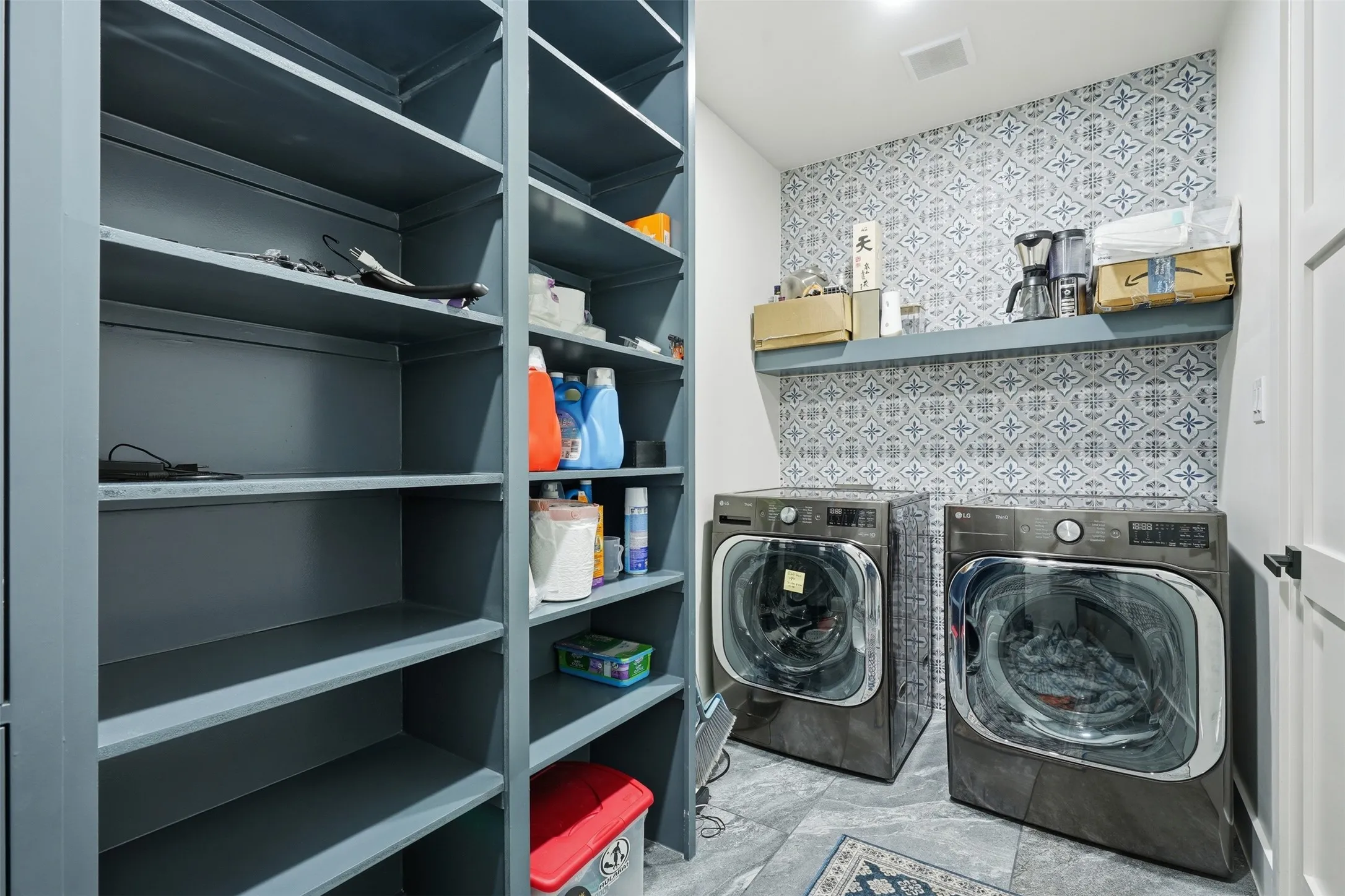 Modern laundry room featuring dual front-load washers with large-capacity glass doors, geometric patterned tile backsplash, open shelving with metal framing for storage and organization, polished concrete flooring, and recessed ceiling lighting