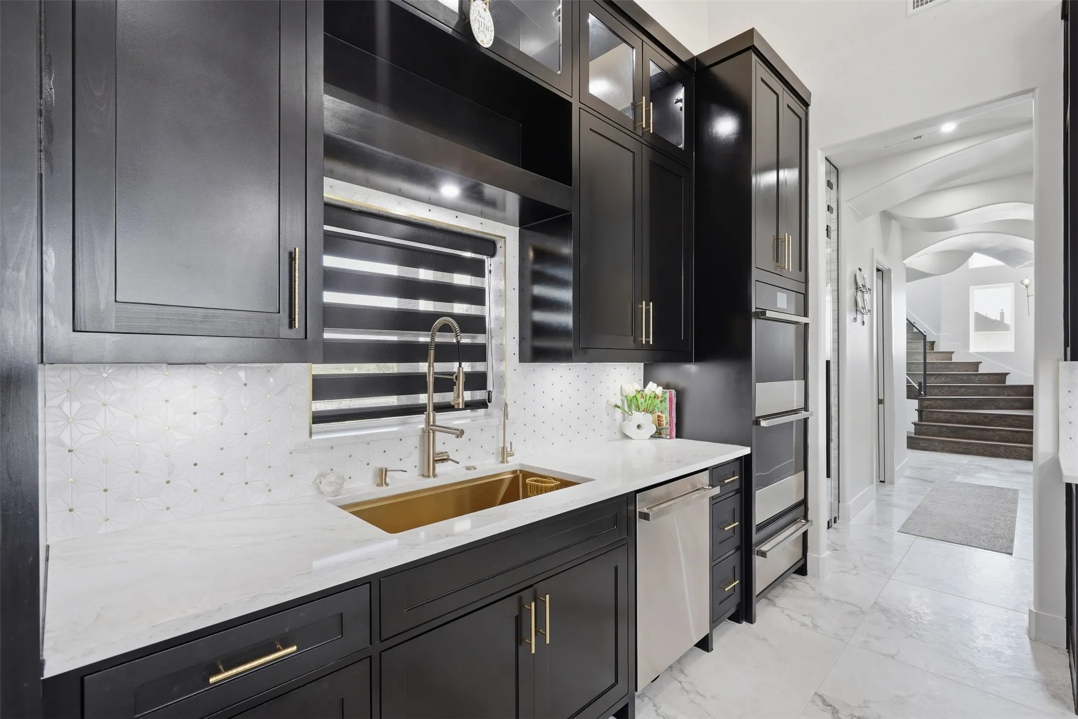 Custom black cabinetry with gold hardware, white countertop, patterned tile backsplash, built-in stainless steel appliances, and a gold-tone sink