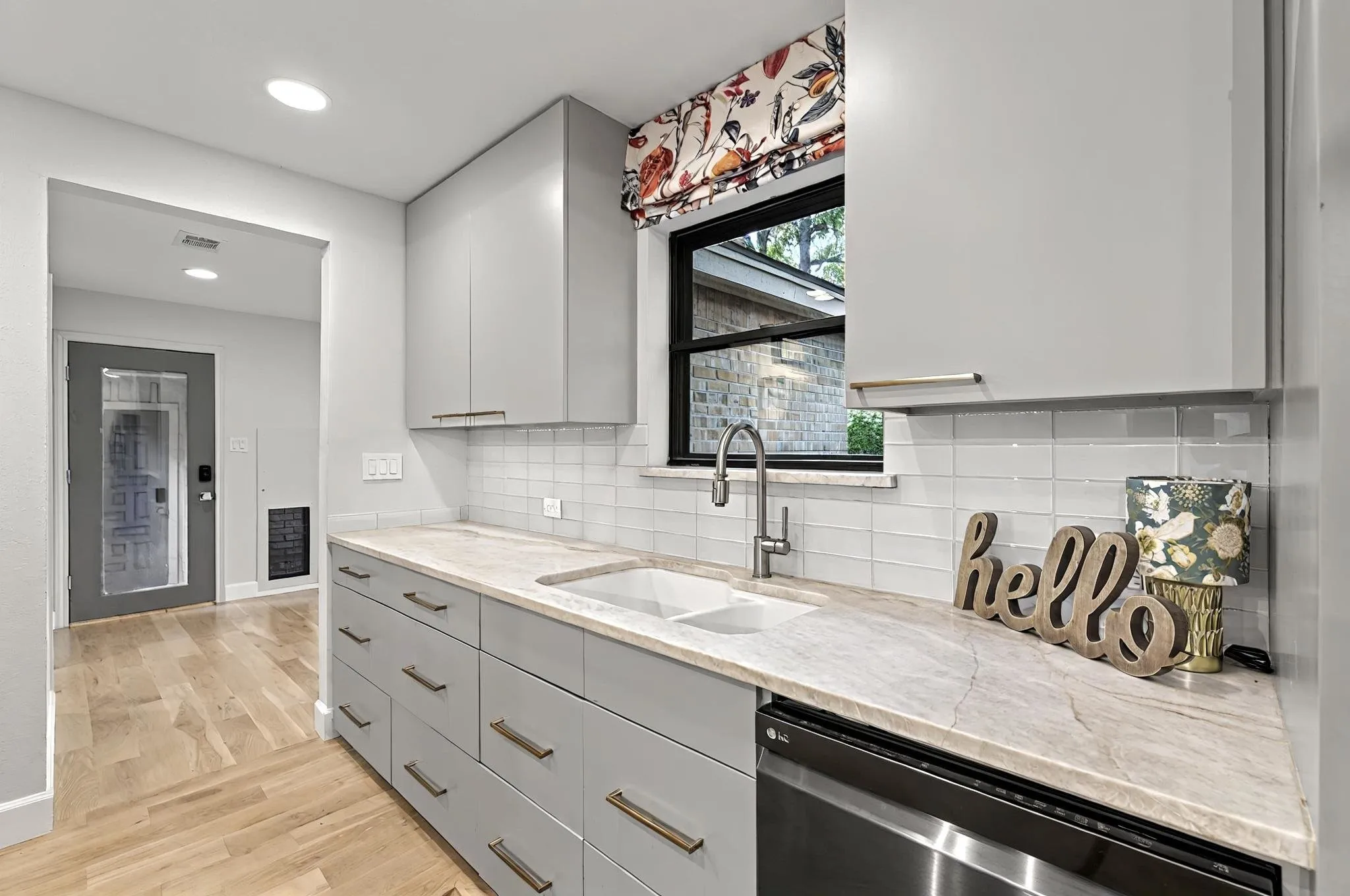 Light wood-finish flooring, light gray cabinetry with gold-tone hardware, a white undermount sink with a gooseneck faucet, light tile backsplash, and a light-toned countertop