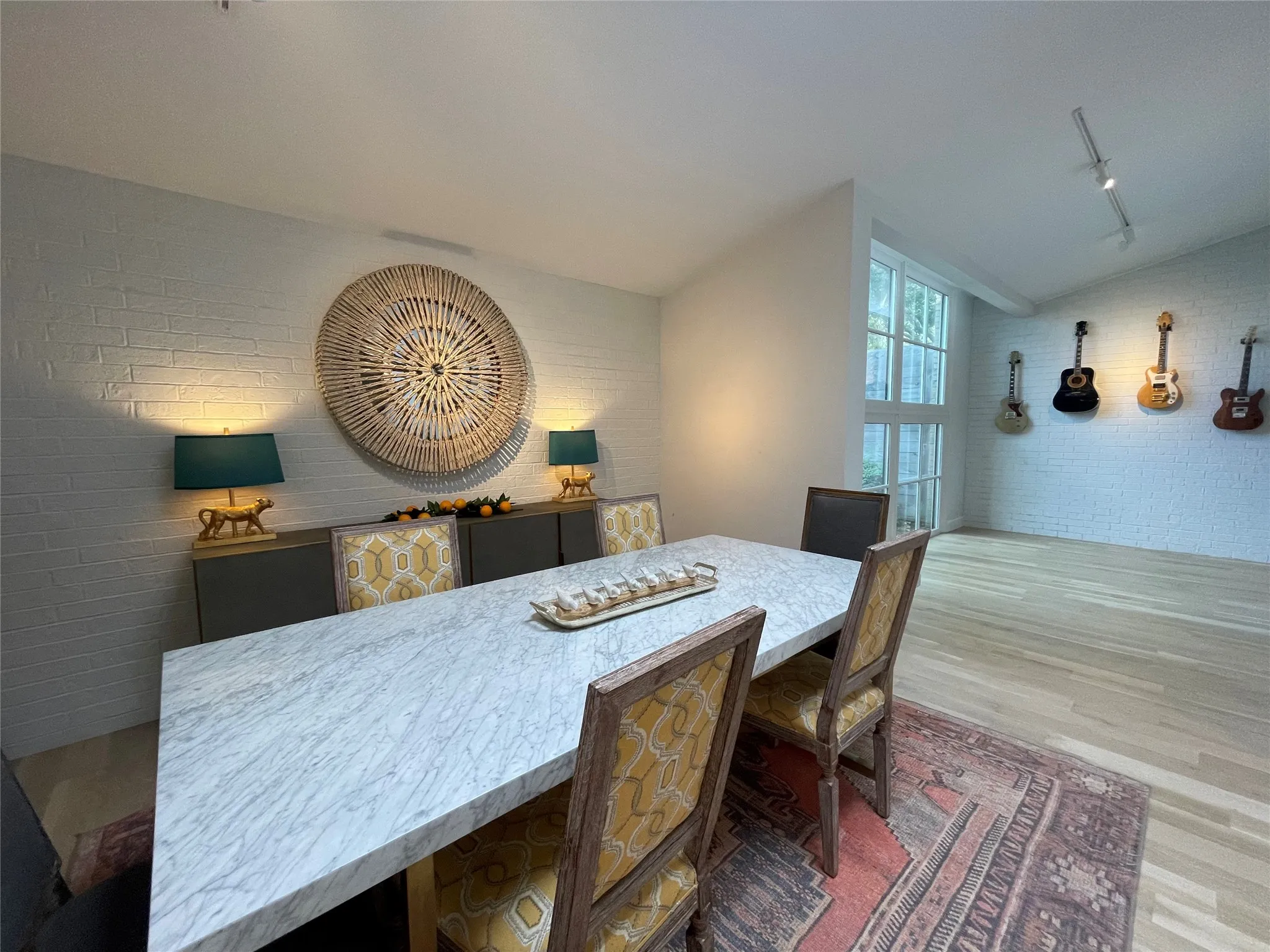 White brick accent wall, high vaulted ceiling, track lighting, large windows, and light wood-finish flooring