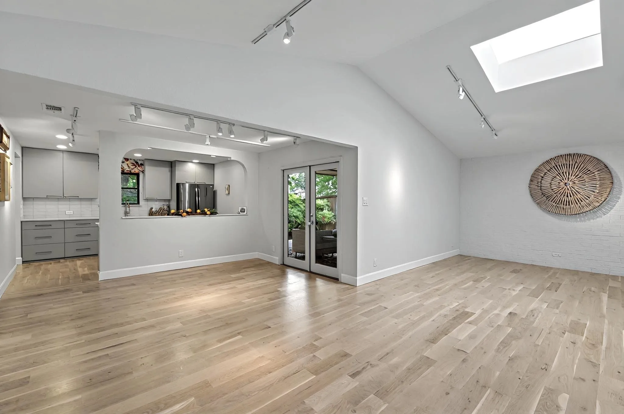 Vaulted ceiling with a skylight, wood-finish flooring, kitchen with gray cabinetry and subway tile backsplash, stainless steel refrigerator, and glass-paned double doors
