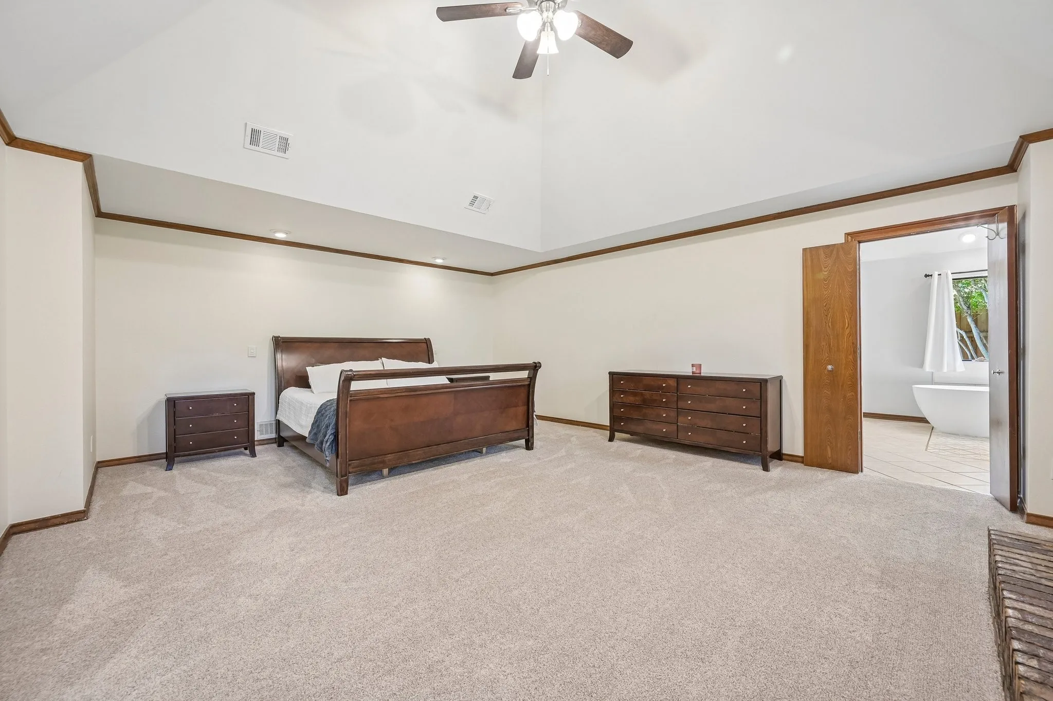 Spacious primary bedroom suite featuring soaring vaulted ceilings with ceiling fan, warm neutral wall finishes accented by wood trim molding, plush carpeting throughout, and ample natural light from multiple windows
