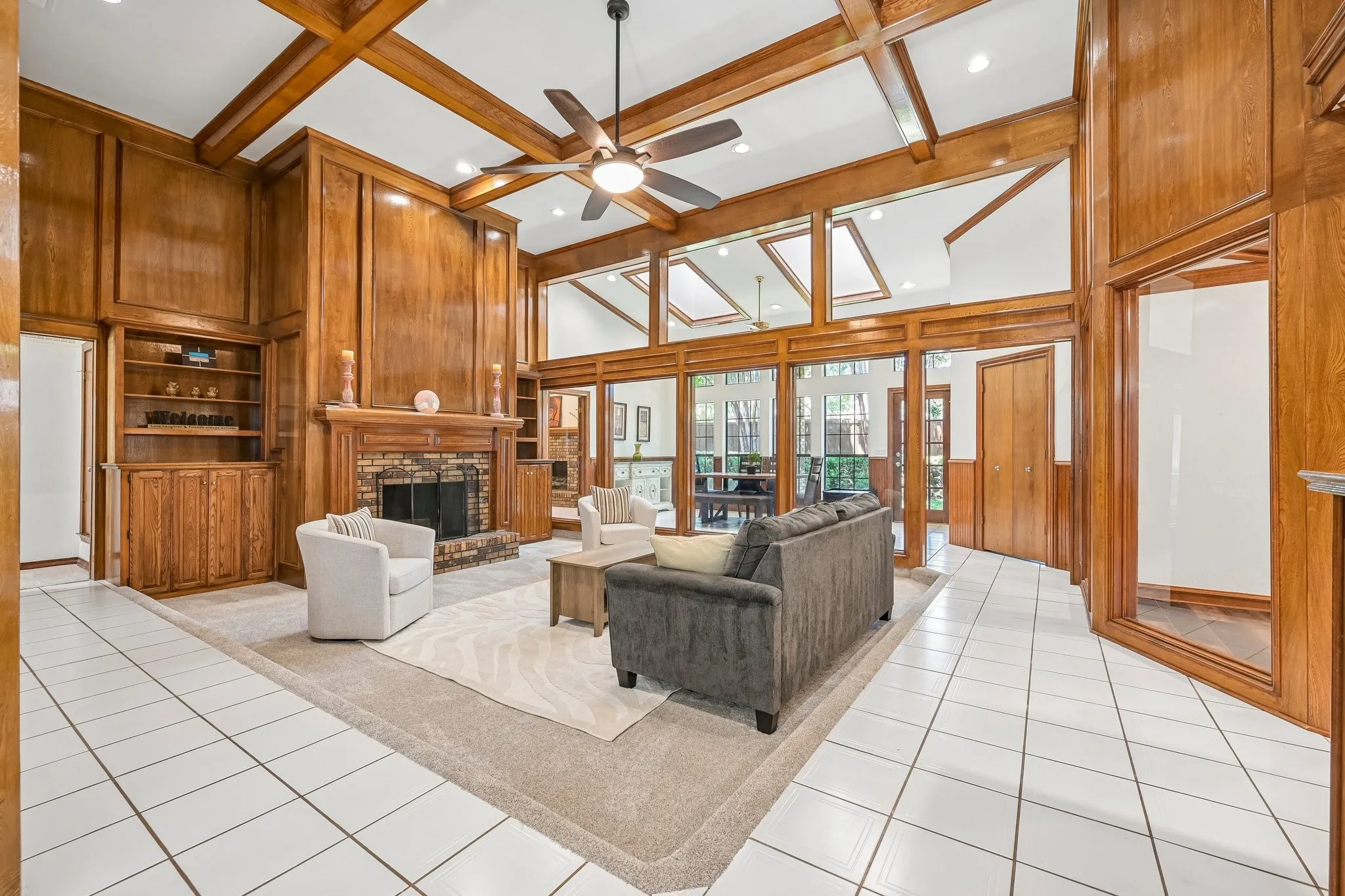 Wood-paneled living space featuring a fireplace with a brick surround, built-in shelving, white tile flooring, and a beamed ceiling