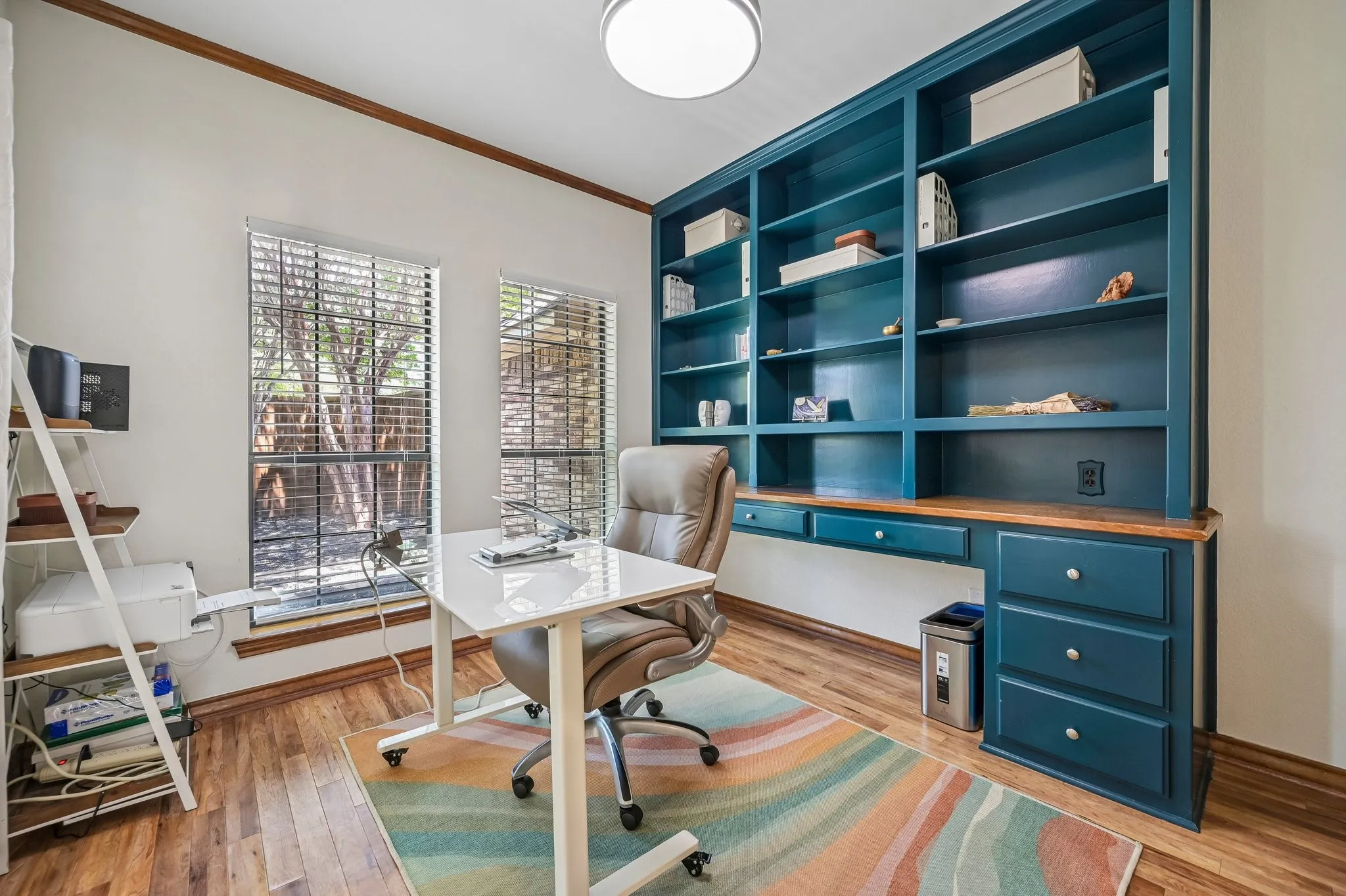 Built-in shelving with integrated desk, hardwood-finish flooring, two window openings with blinds, and recessed lighting