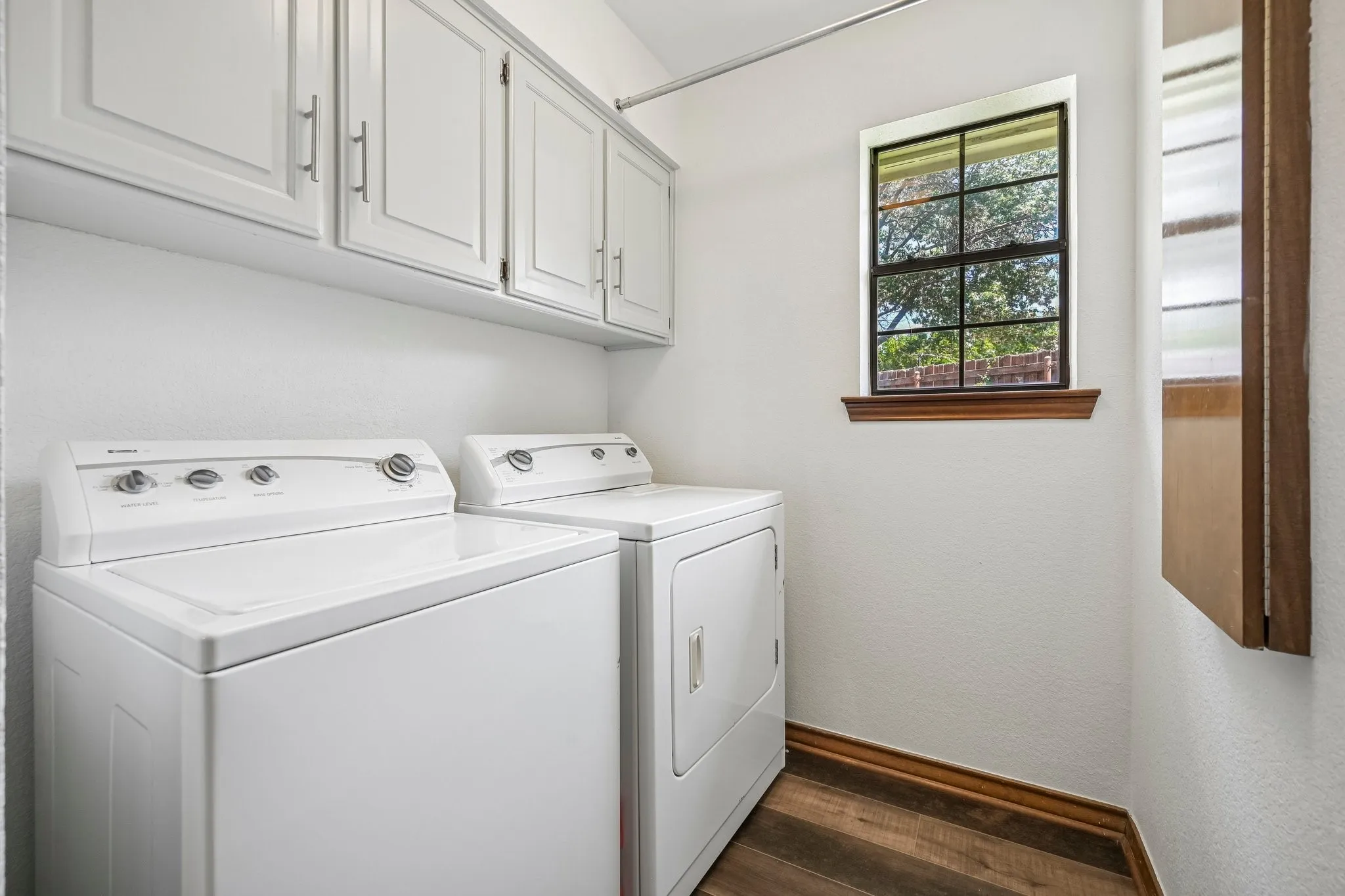 White shaker-style cabinetry, wood-finish flooring, and a single-pane window with wood trim