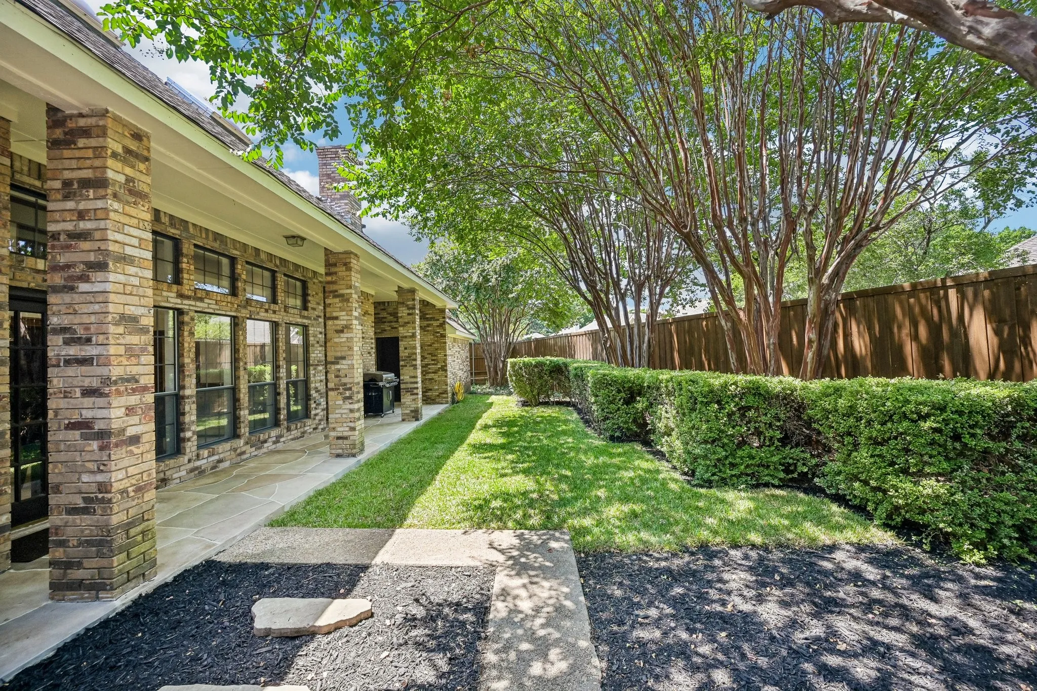 Brick exterior with expansive backyard featuring manicured lawn, mature shade trees, and mature hedge landscaping