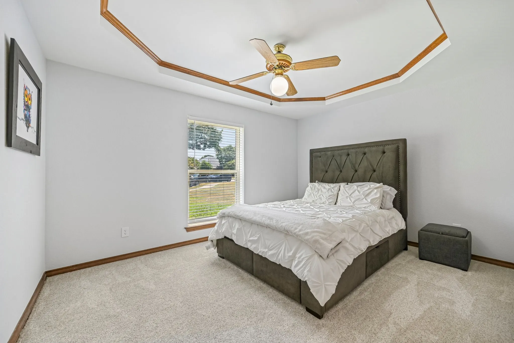 Spacious primary bedroom with wood-trim architectural ceiling detail, bronze ceiling fan with integrated lighting, upholstered headboard bed frame, and large window with pastoral landscape backdrop