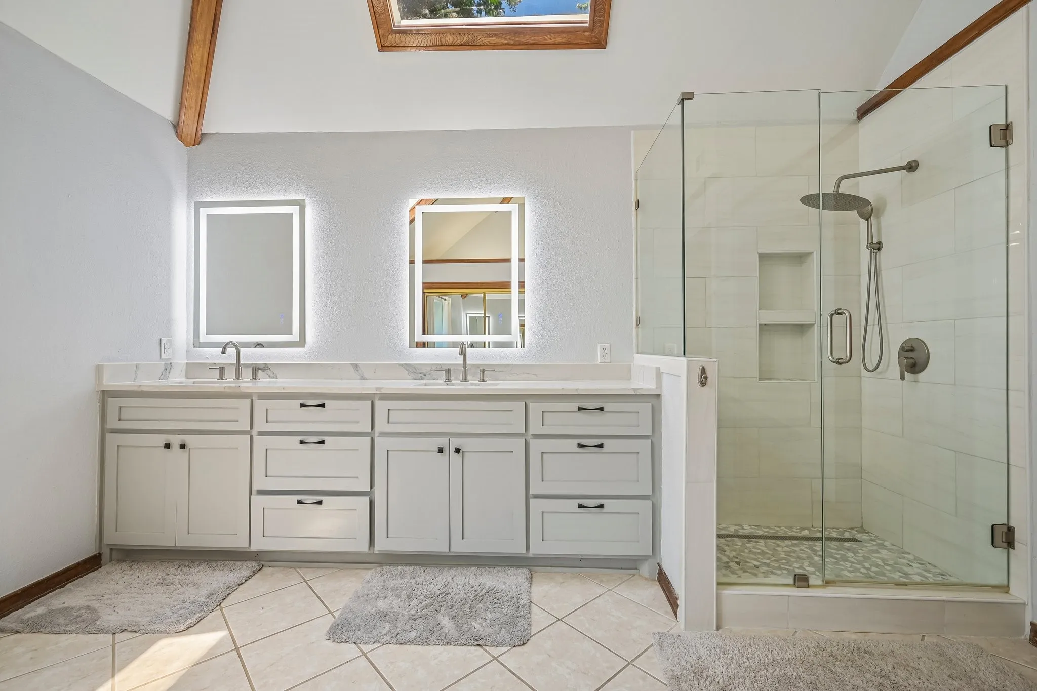 Vaulted ceiling with exposed wood beams, skylight, dual vanity with undermount sinks and quartz countertops, illuminated mirrors, and a glass-enclosed shower with tiled walls and a rain shower head