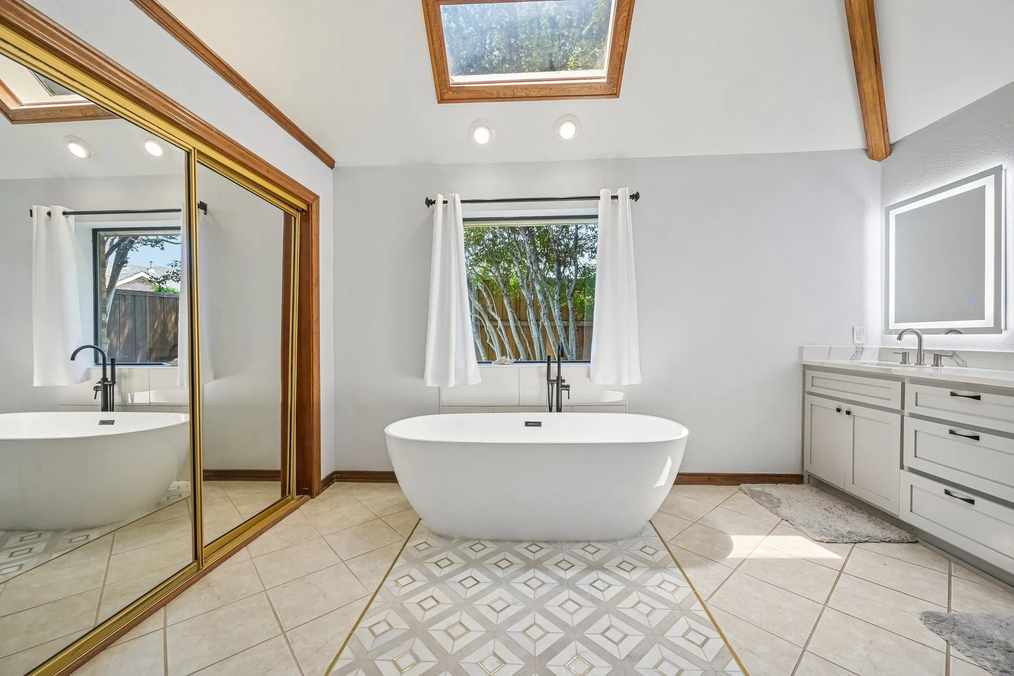 Freestanding soaking tub with a black faucet, dual vanity with a lighted mirror, skylight, and large mirrored closet doors