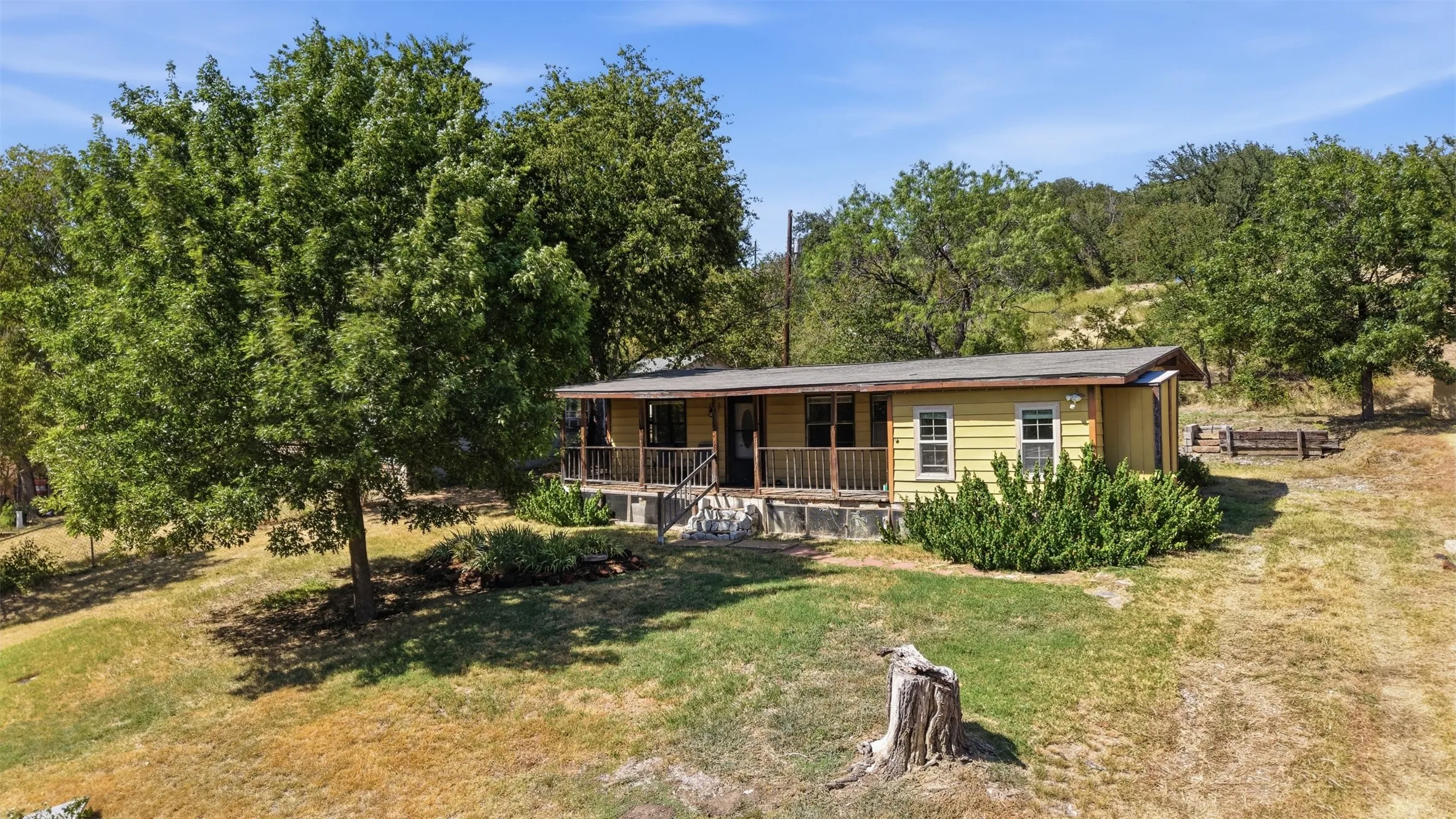 Yellow exterior with a covered porch featuring wood railings, set on a sloped landscape with mature trees and natural ground cover