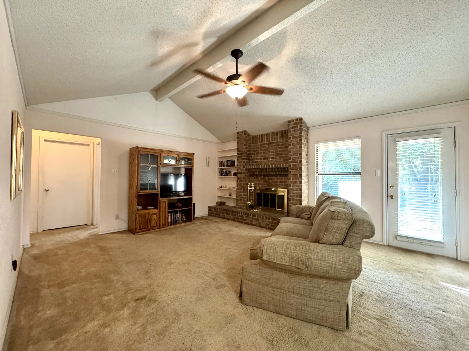 Living Room: brick WOOD-Burning Fireplace, built-in shelving, vaulted ceiling with ceiling fan, and carpeted flooring. Back door opens to covered patio.