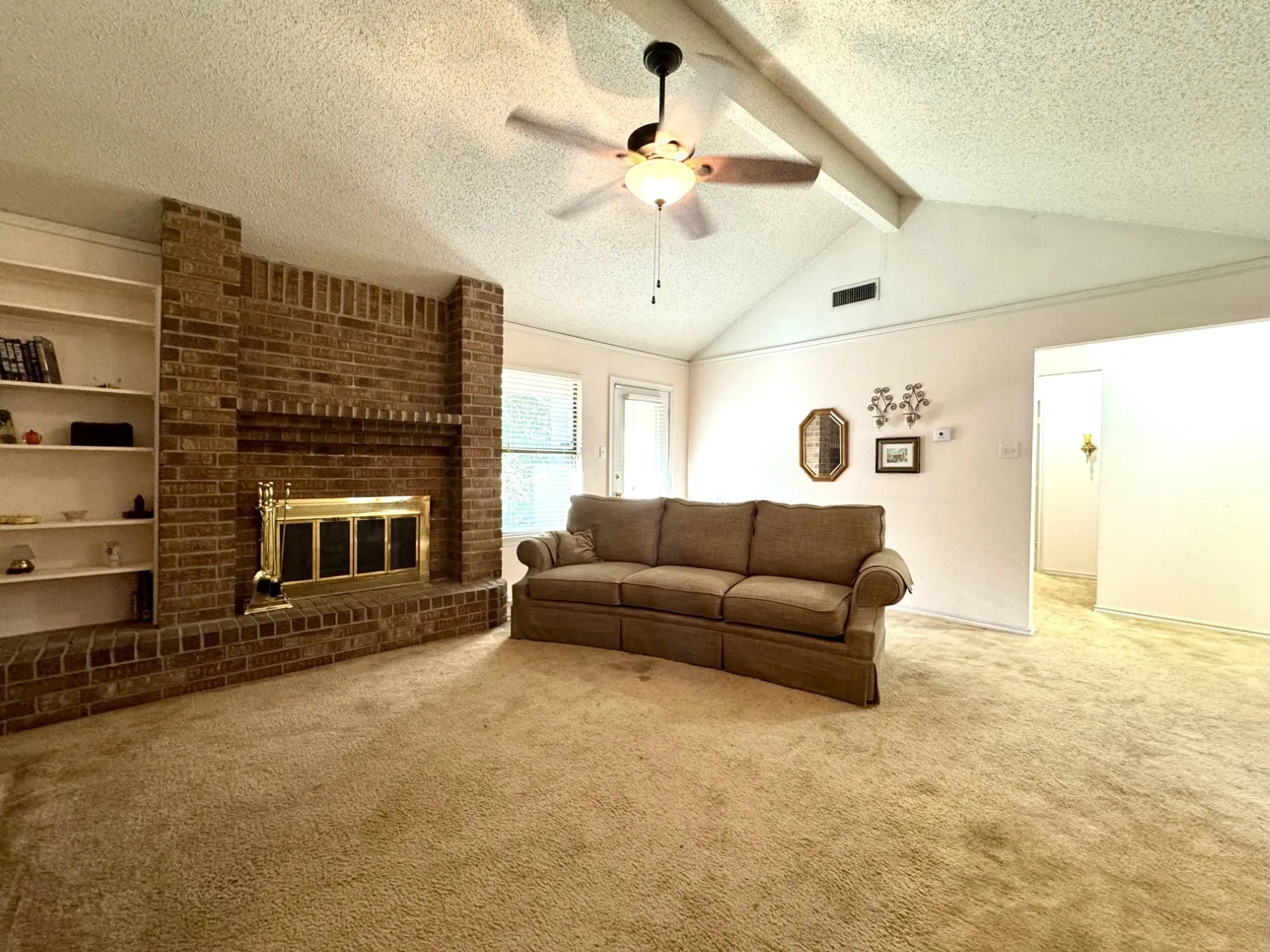 Spacious Living Room features a brick wood-burning fireplace with glass & brass door, built-in shelving, vaulted ceiling with fan, and carpeted flooring.