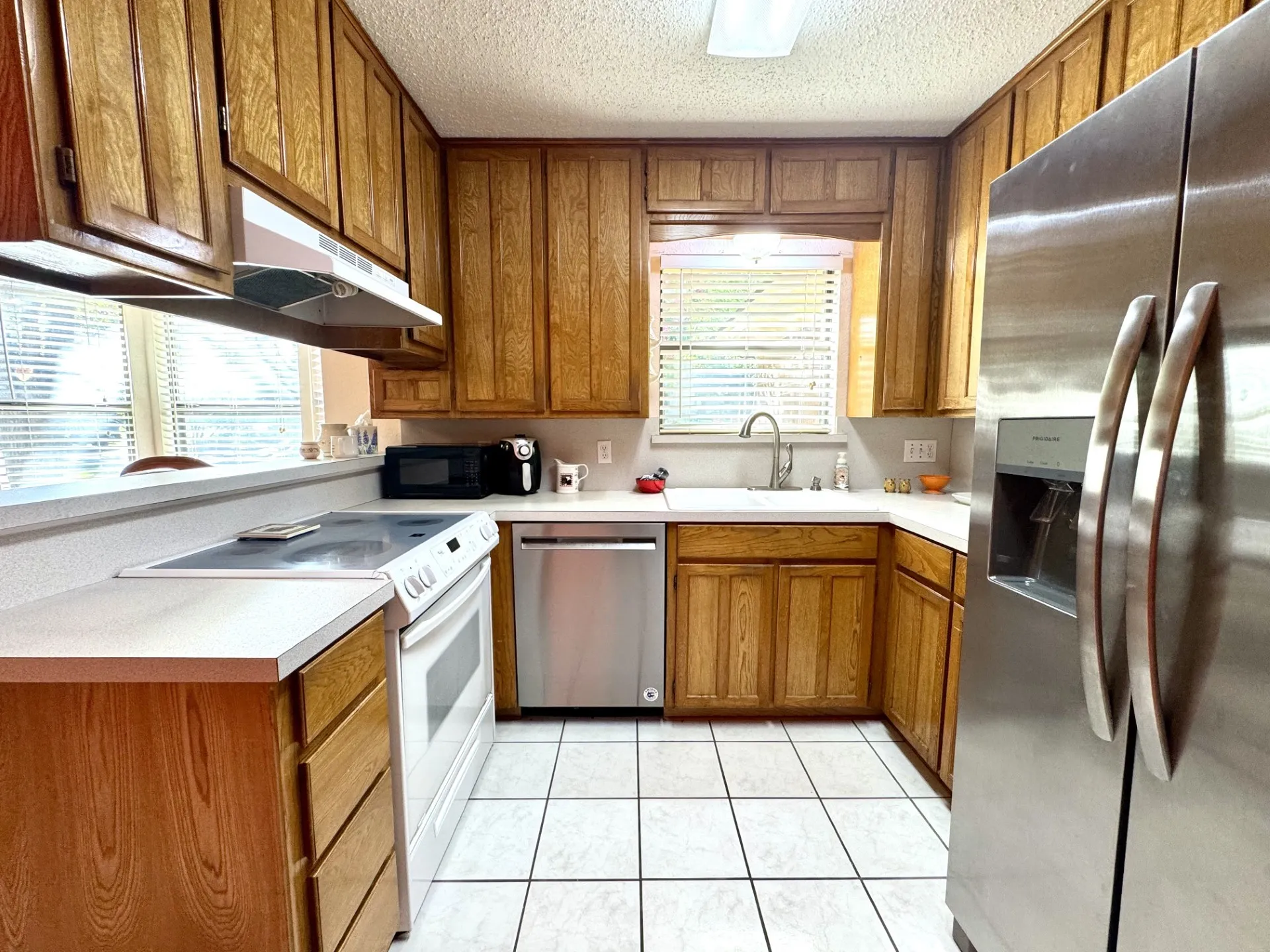 Kitchen features double sink, wood cabinetry, white countertops, white tile flooring, white electric range, microwave, stainless steel dishwasher and refrigerator.
