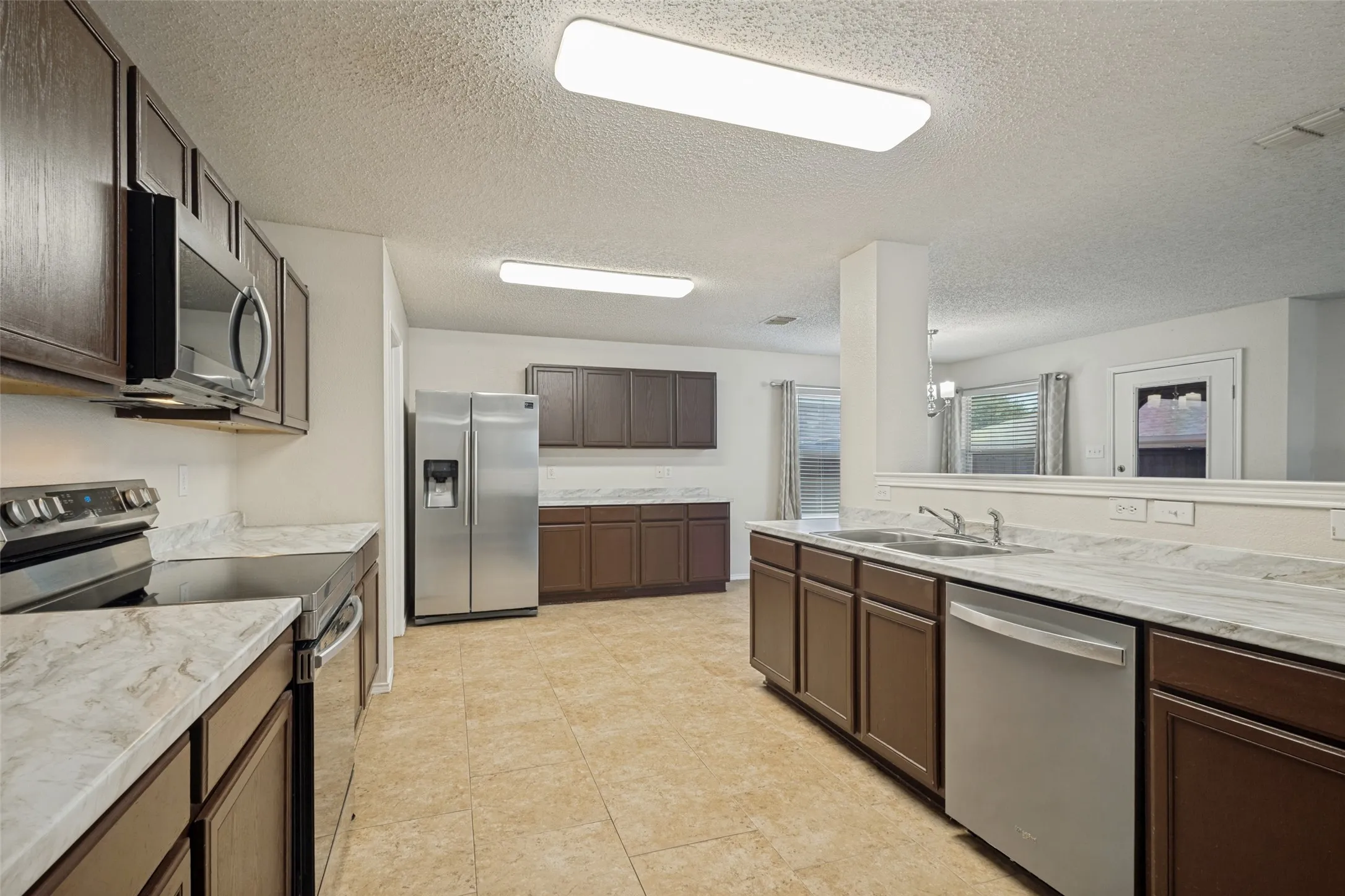 Galley-style kitchen featuring stainless steel appliances, dual sink stations with marble-finish countertops, dark wood cabinetry, and ceramic tile flooring throughout