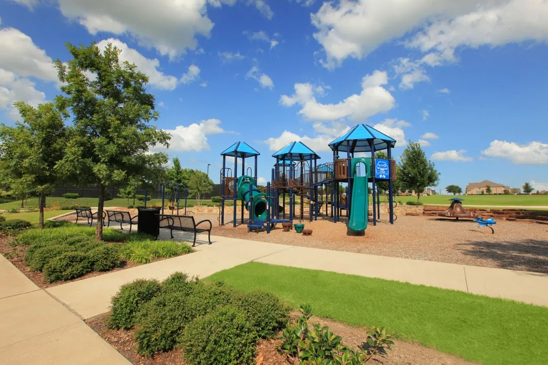 Community park featuring a multi-level playground structure with blue architectural accents, rubberized safety surfacing, manicured lawn areas, mature shade trees, park benches, and paved walking paths throughout the grounds