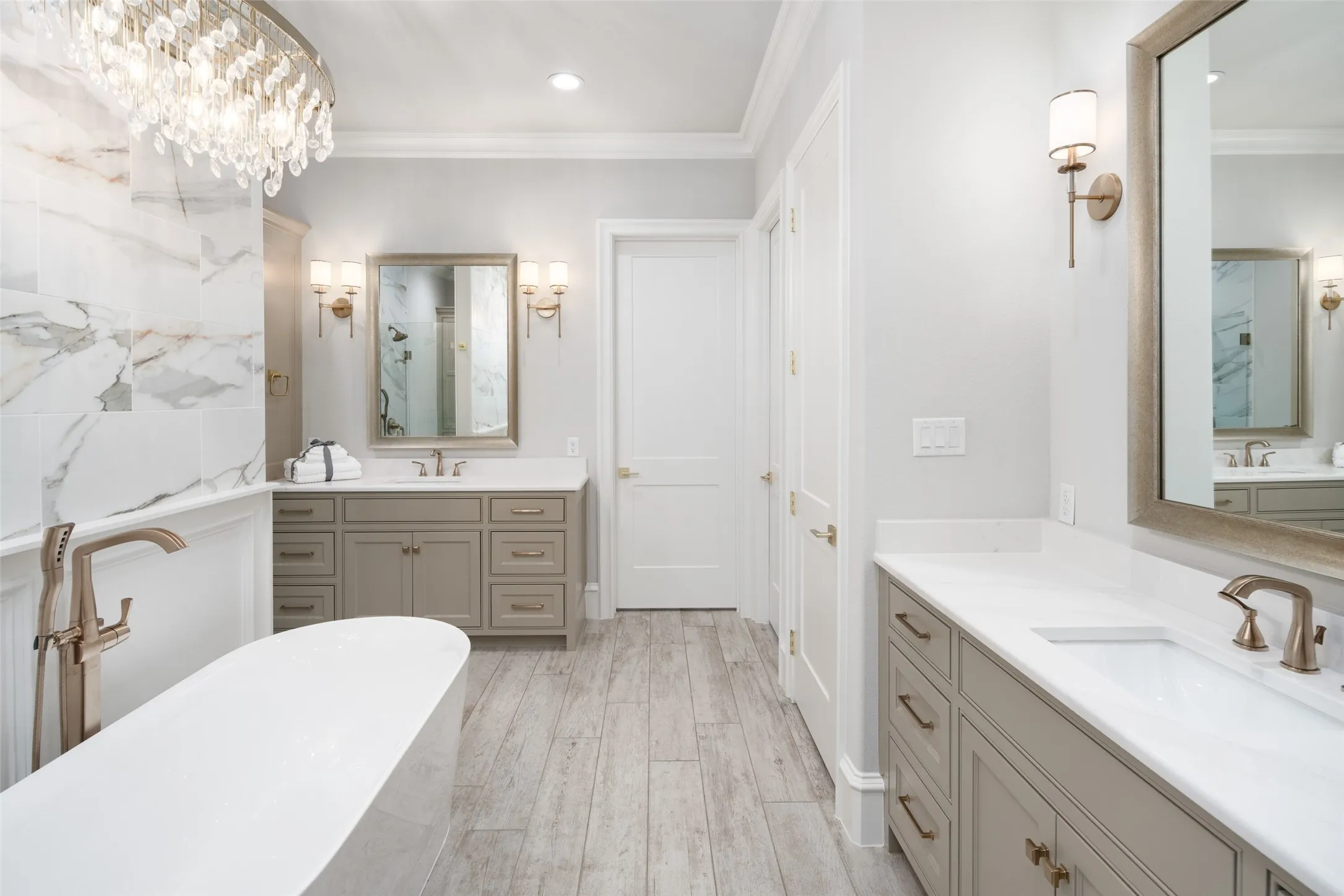 Freestanding soaking tub with floor-mounted faucet, dual vanities with integrated sinks and white countertops, wood-finish flooring, and a crystal chandelier