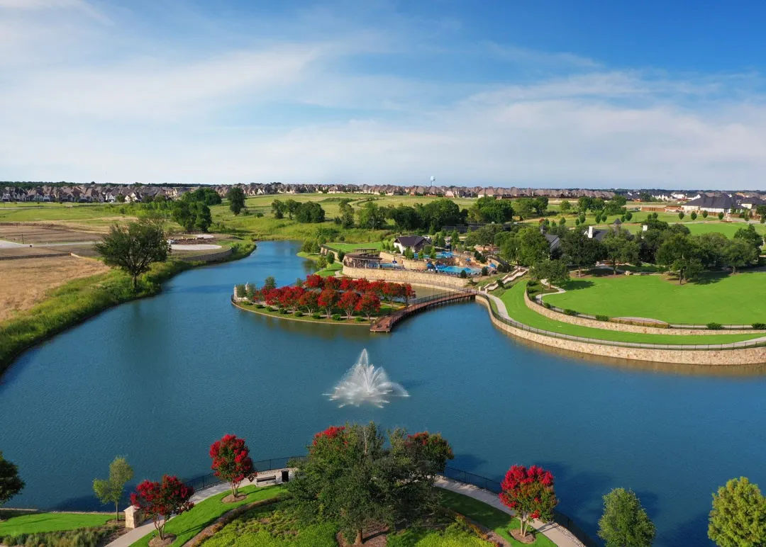 Aerial view showcasing manicured grounds with verdant landscaping, a pristine blue-water pond featuring a central fountain, wooden pedestrian bridges, ornamental plantings with seasonal color accents, and mature tree specimens throughout the property