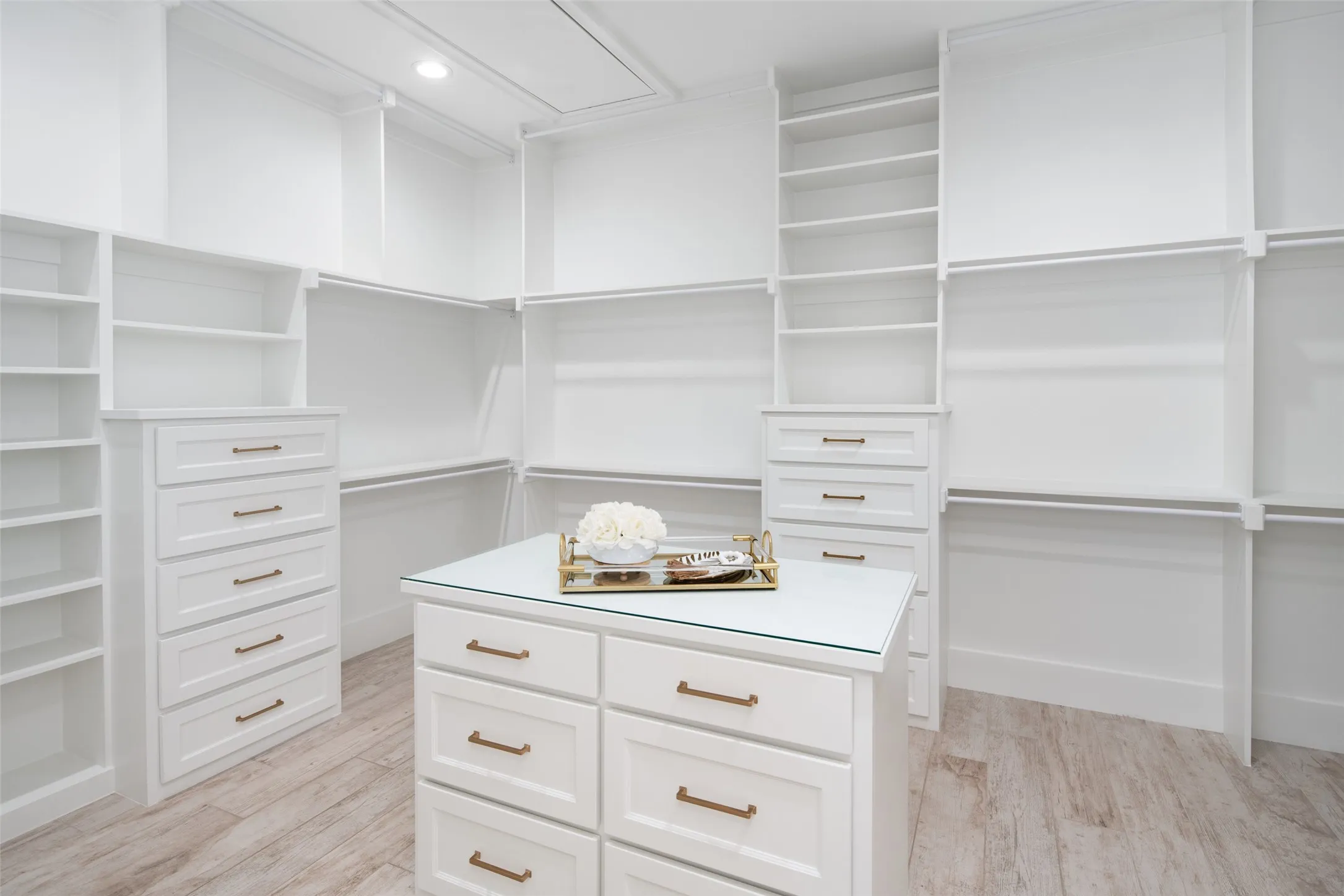 Spacious custom closet featuring white built-in cabinetry, integrated shelving, ample hanging rods, a central island with glass top, and wood-finish flooring