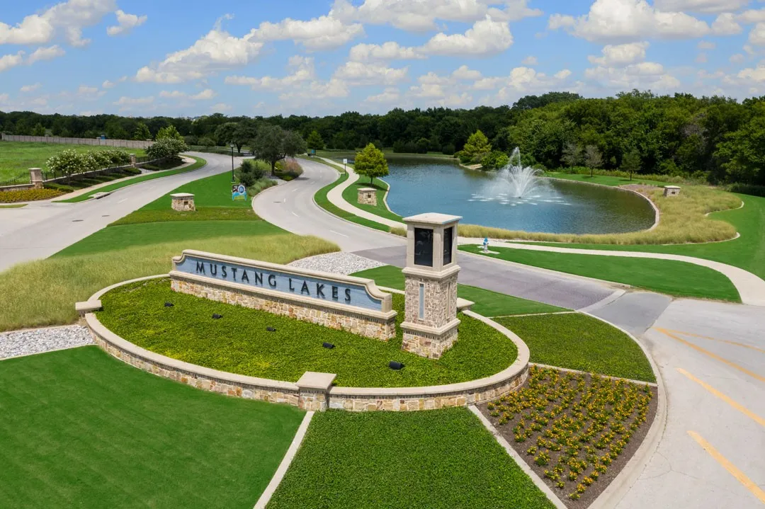 Mustang Lakes community entrance featuring a professionally landscaped median with mature trees, manicured lawn, and decorative hardscaping
