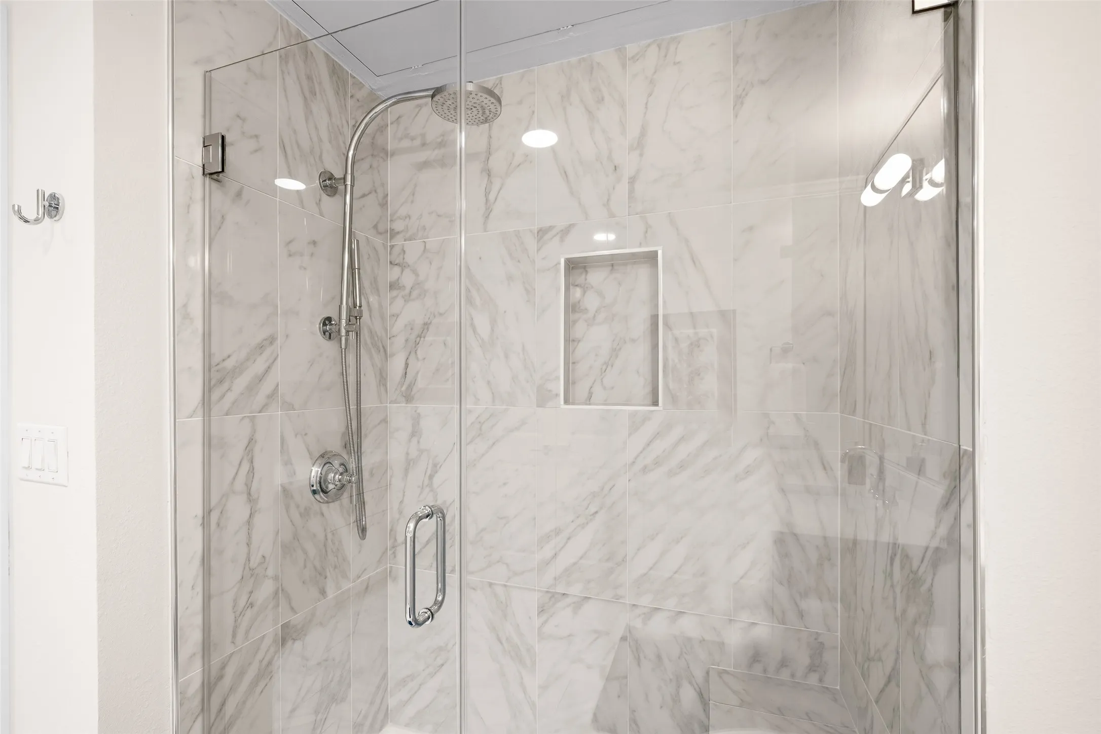 Glass-enclosed shower with wall-mounted rainfall showerhead, handheld shower fixture, built-in shelving, and light-toned tile surround