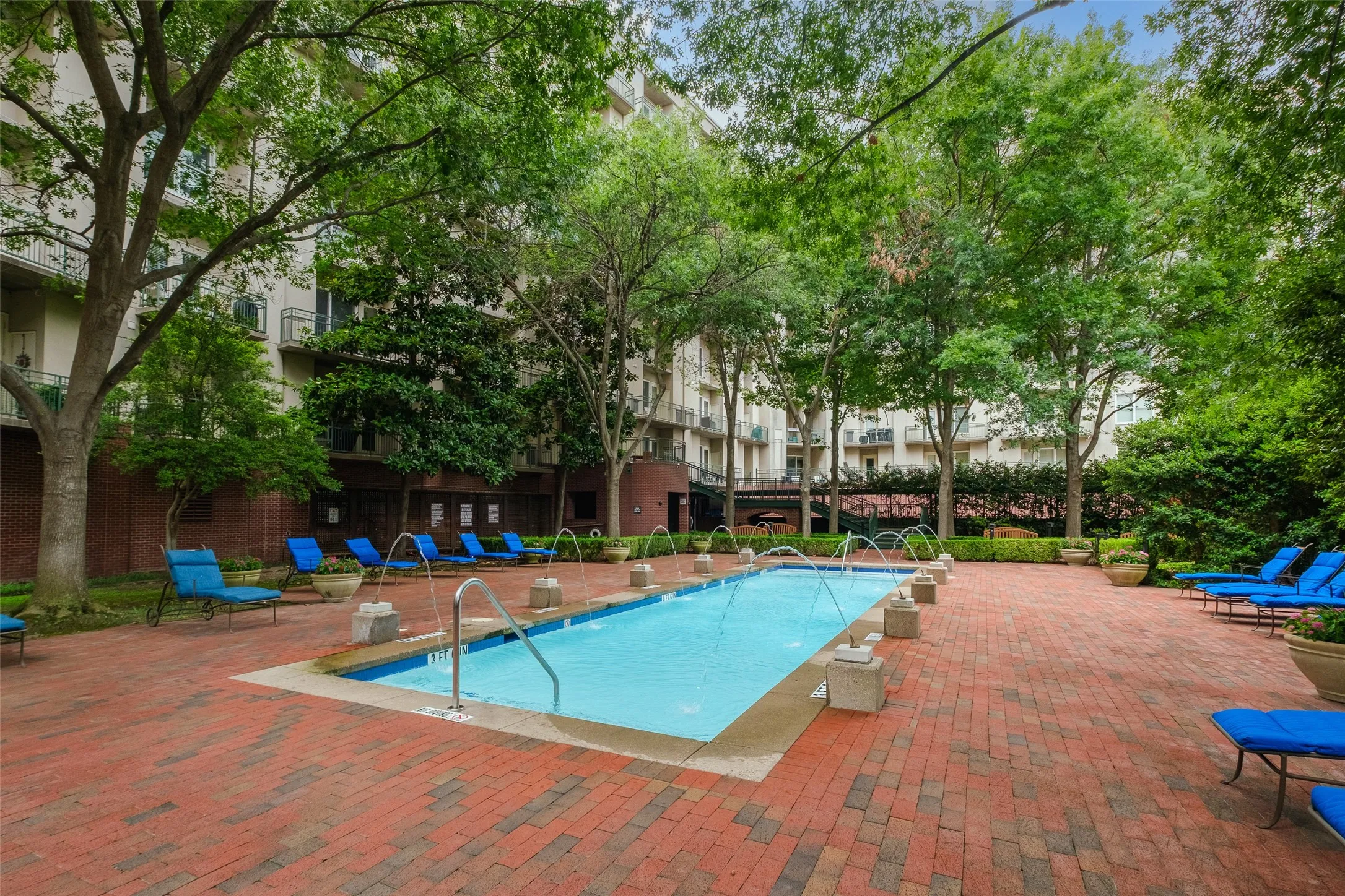 Outdoor swimming pool with integrated water features, surrounded by brick paving and mature trees