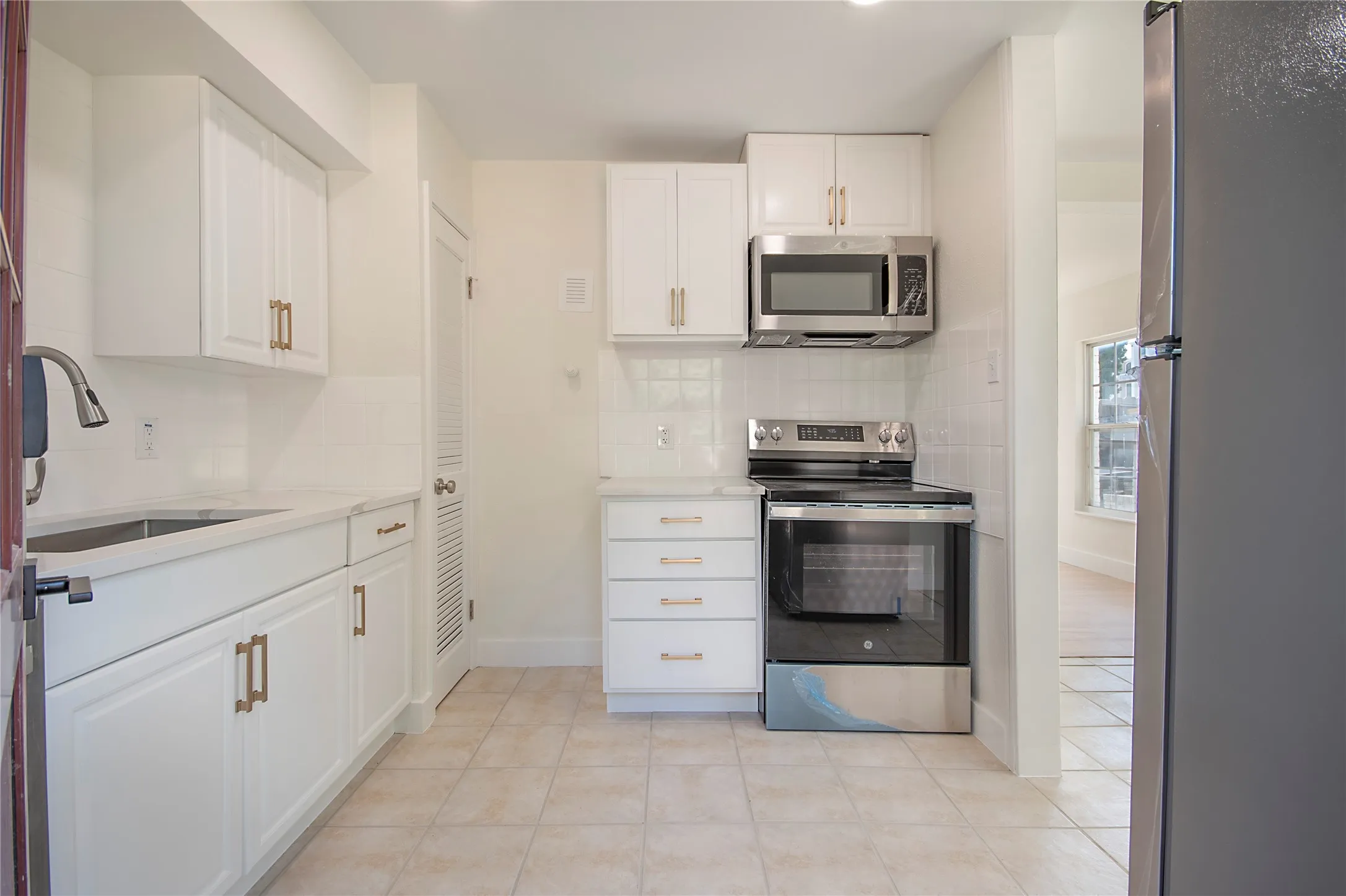 White shaker-style cabinetry with gold-tone hardware, stainless steel appliances, white subway tile backsplash, light-toned countertop, and beige tile flooring