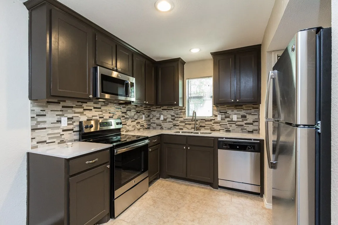 Modern kitchen featuring dark cabinetry with recessed panel detailing, stainless steel appliances including a refrigerator and range, a geometric tile backsplash in neutral tones, quartz countertops, and tile flooring throughout