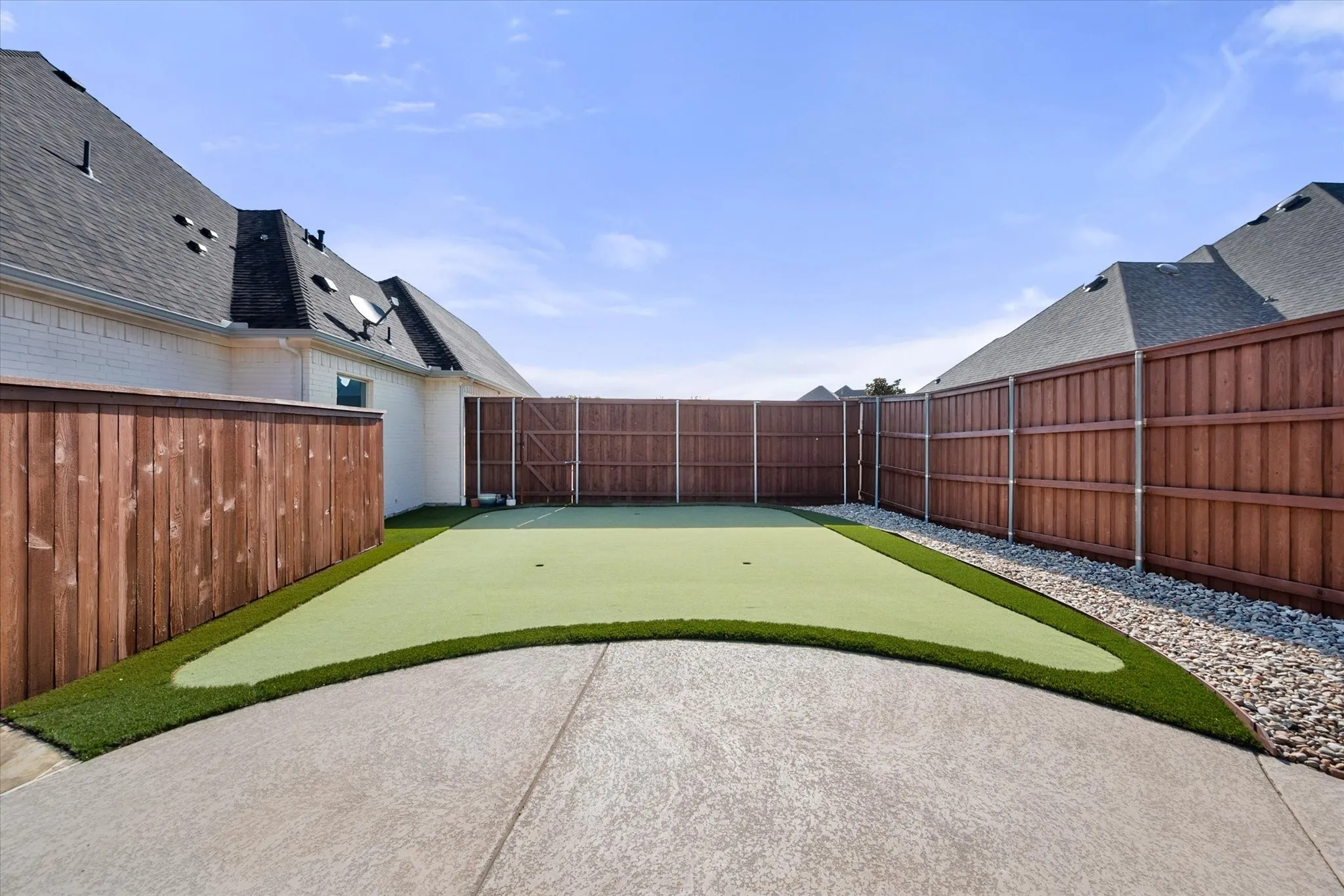 Expansive backyard featuring a putting green with curved design, concrete patio, decorative gravel borders, and mature landscaping framed by wood-finish privacy fencing