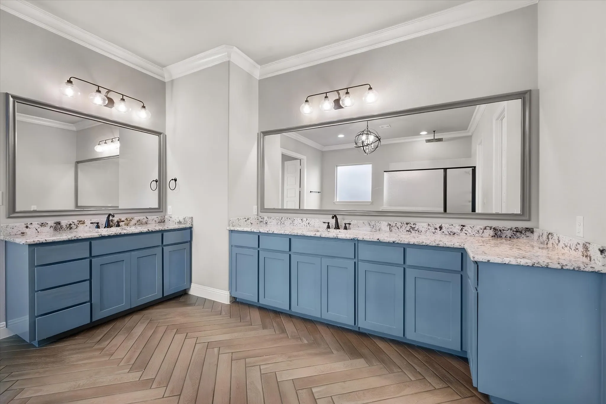 Luxurious dual-vanity bathroom featuring custom blue cabinetry with ample storage, granite countertops, framed mirrors with integrated lighting, and herringbone wood-finish flooring throughout
