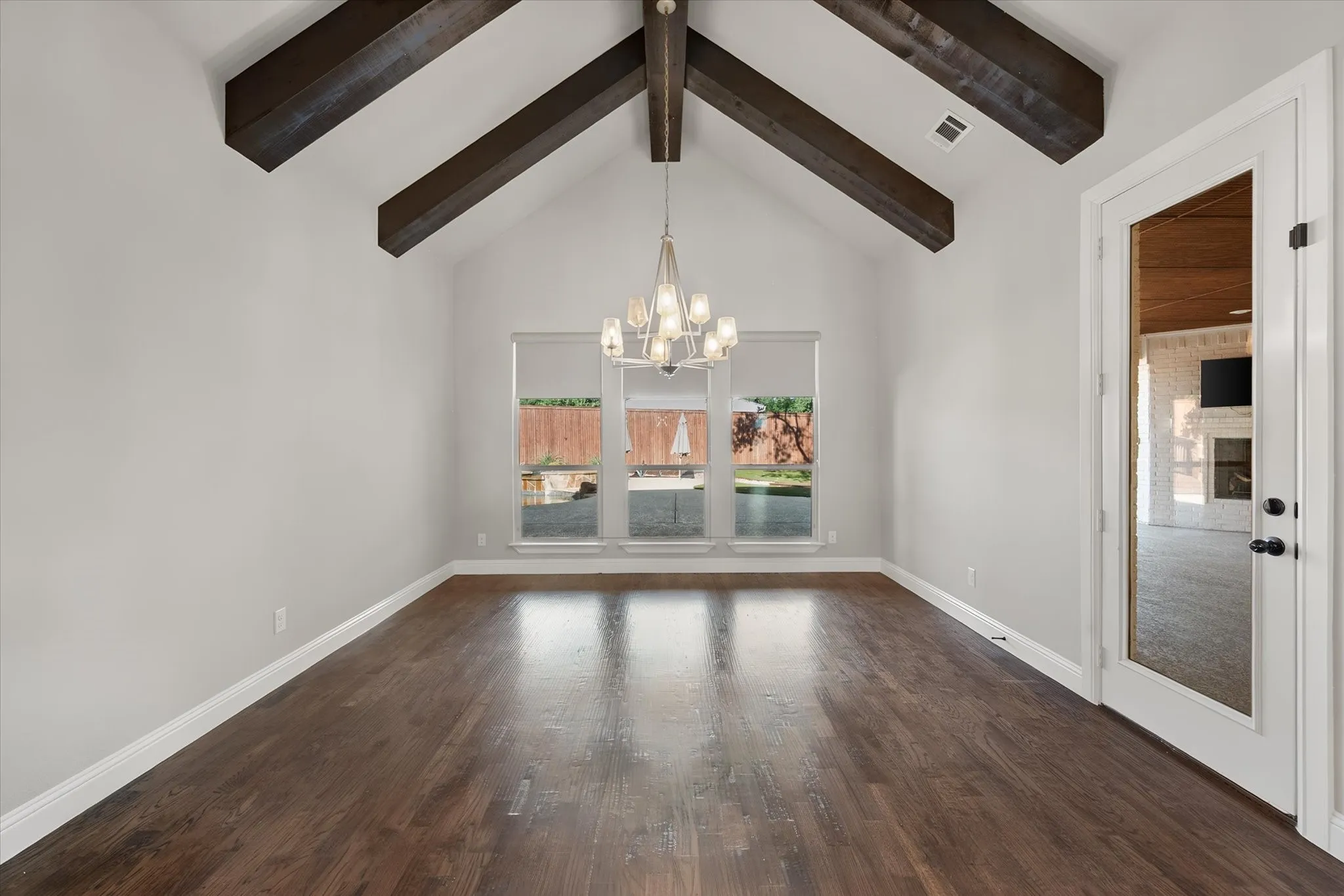 Spacious room featuring exposed wooden ceiling beams, white vaulted ceilings, dark wood-finish flooring, a multi-pane window wall with natural light, and a chandelier fixture suspended from the center beam
