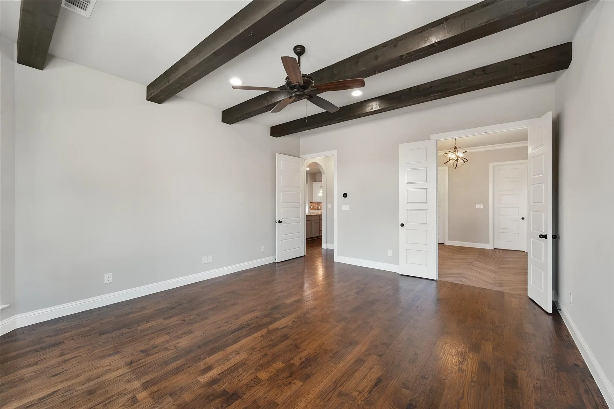 Spacious room featuring wood-finish flooring, exposed ceiling beams, a ceiling fan with integrated lighting, and multiple paneled doors