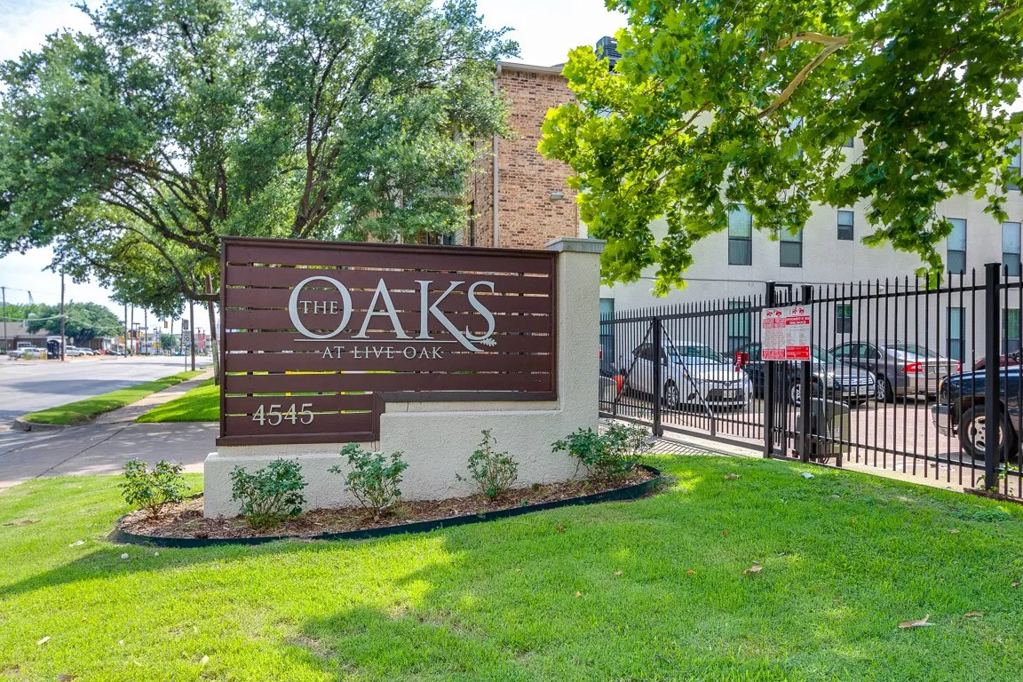 The Oaks at Live Oak community entrance features a distinctive wooden monument sign with bronze lettering, anchoring a manicured landscape of lush green lawn and mature shade trees