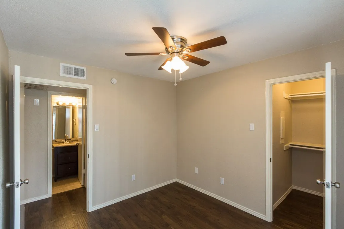 Spacious primary bedroom with wood-finish flooring, ceiling fan with integrated lighting, dual walk-in closets with built-in shelving and hanging systems, and an ensuite bathroom featuring a vanity with ample counter space and warm ambient lighting