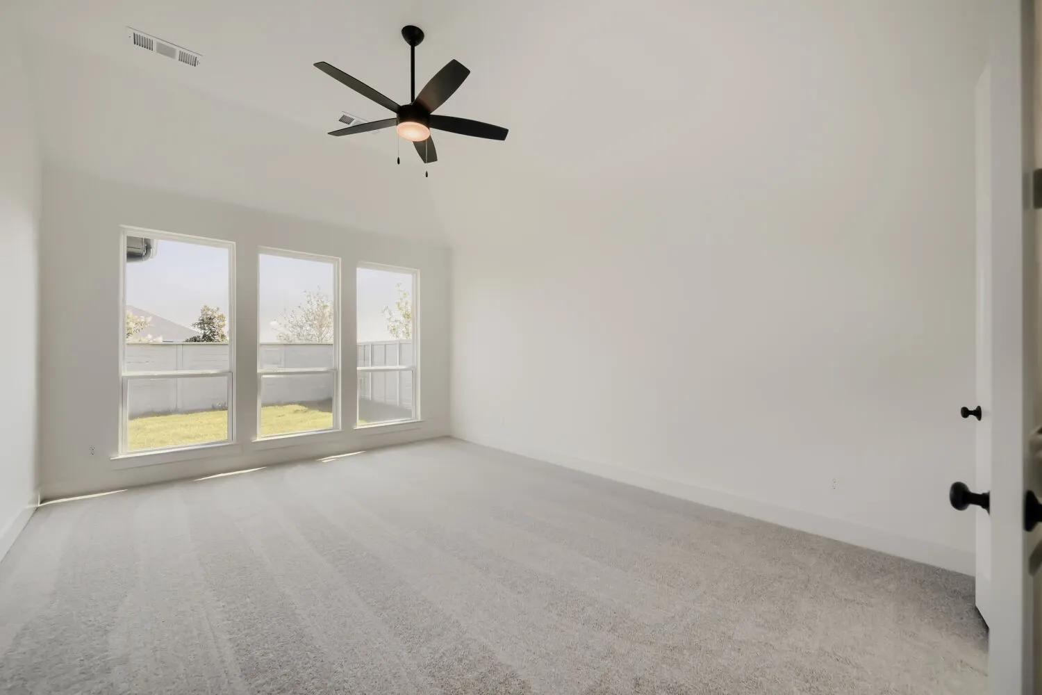Vaulted ceiling, three large windows, dark-finish ceiling fan, light gray carpet, and white interior door with dark hardware