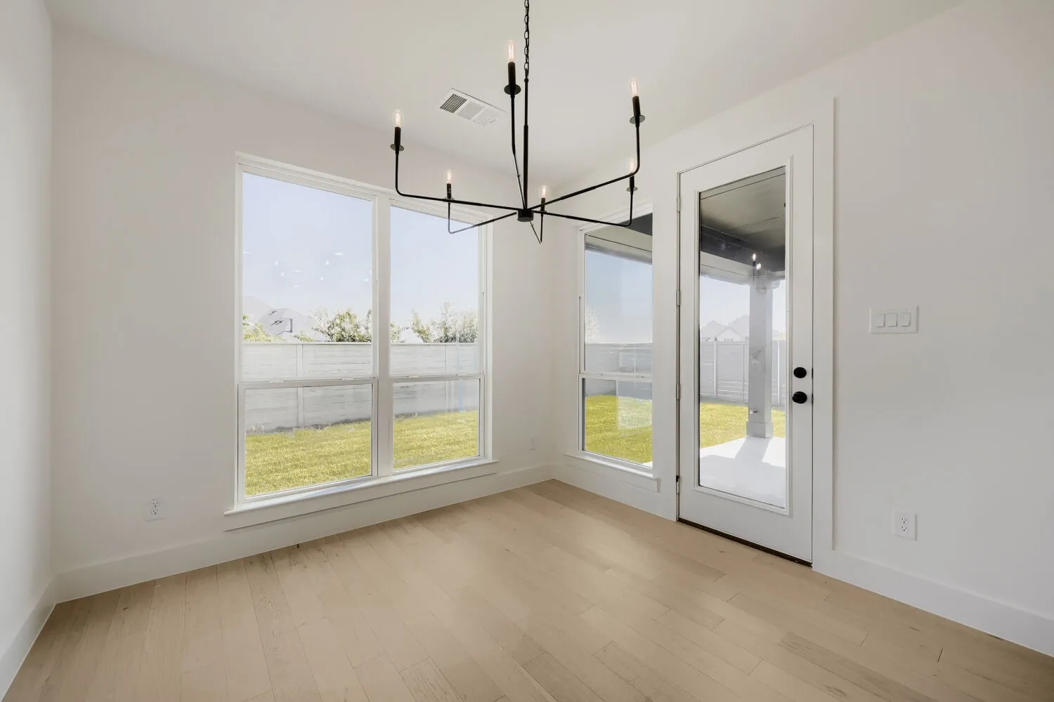 Light wood-finish flooring, a modern black chandelier, white walls, and a full-light glass door leading to an outdoor patio