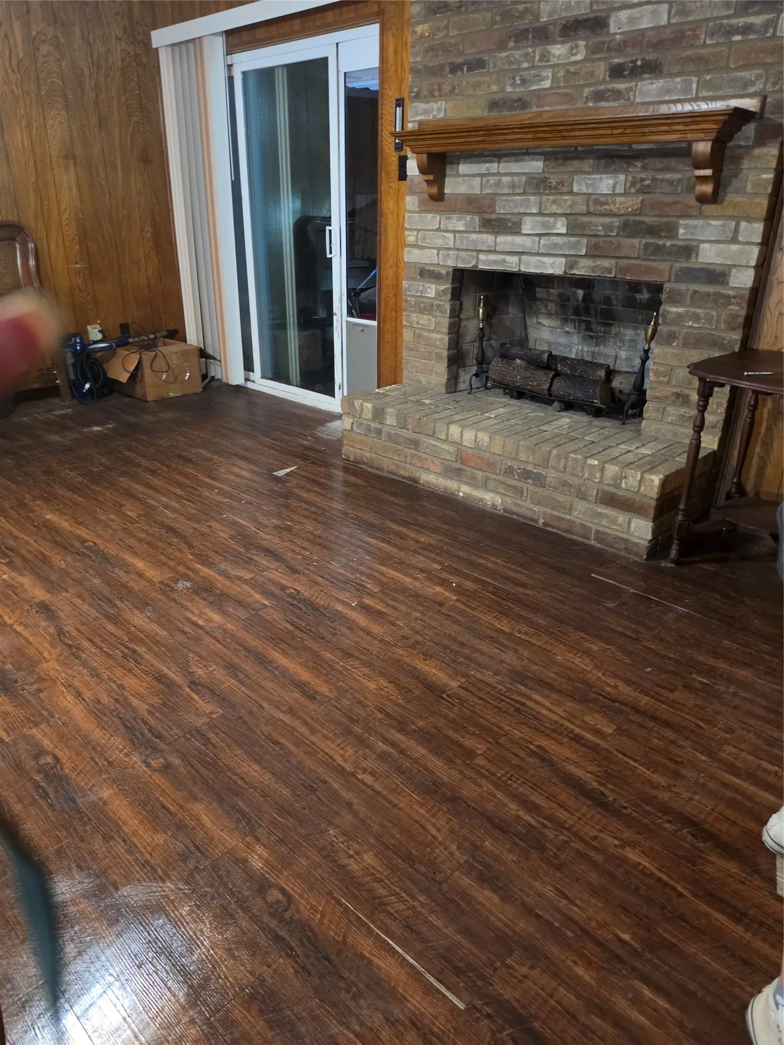 Spacious living area showcasing rich wood-finish flooring throughout, brick fireplace with wooden mantel, and glass sliding doors leading to outdoor space